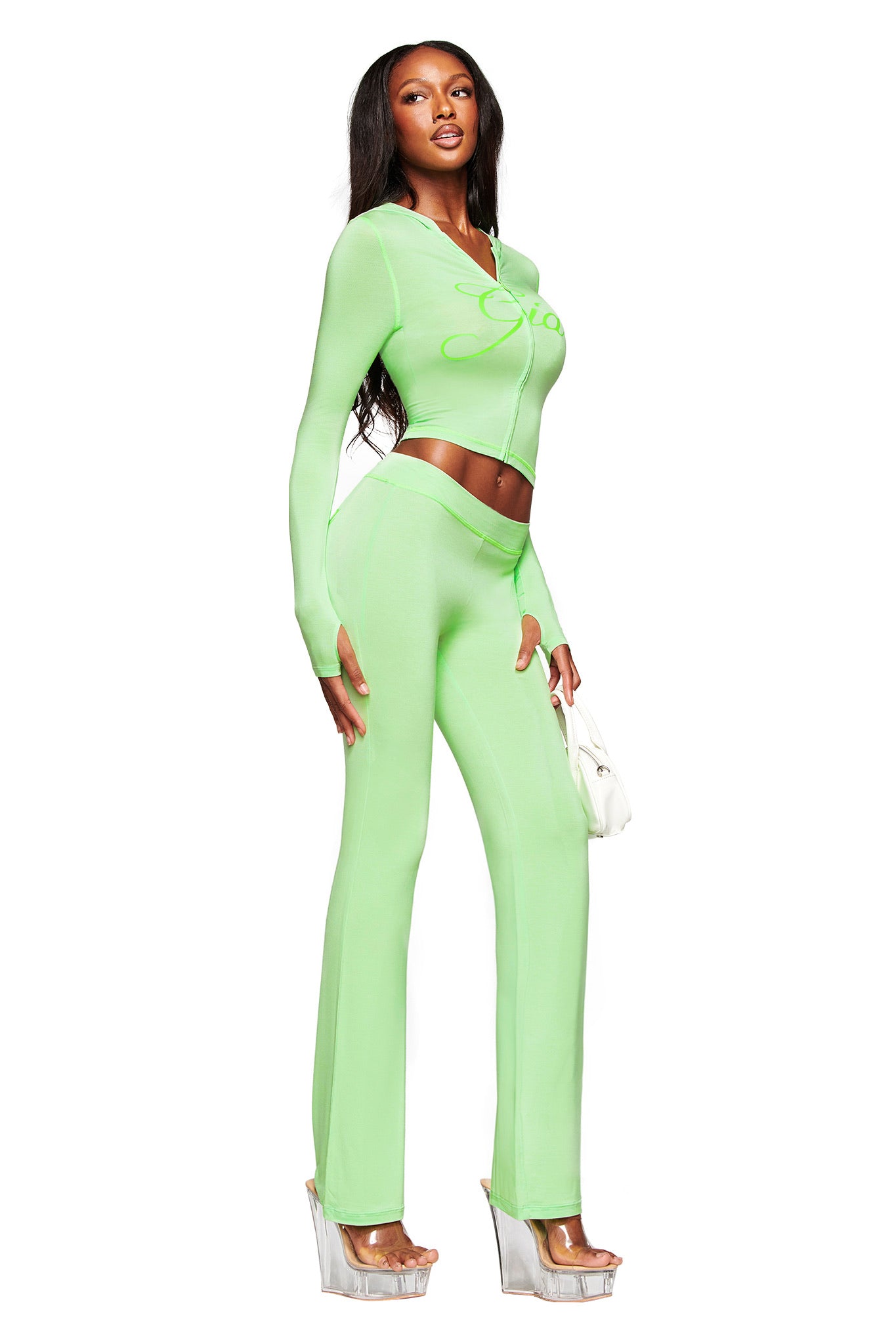 A woman models the I.AM.GIA BLARE TRACKPANT in neon green, a set with a long-sleeve crop top and high-waisted, flared pants. She pairs it with a white handbag and clear platform heels against a plain white background.