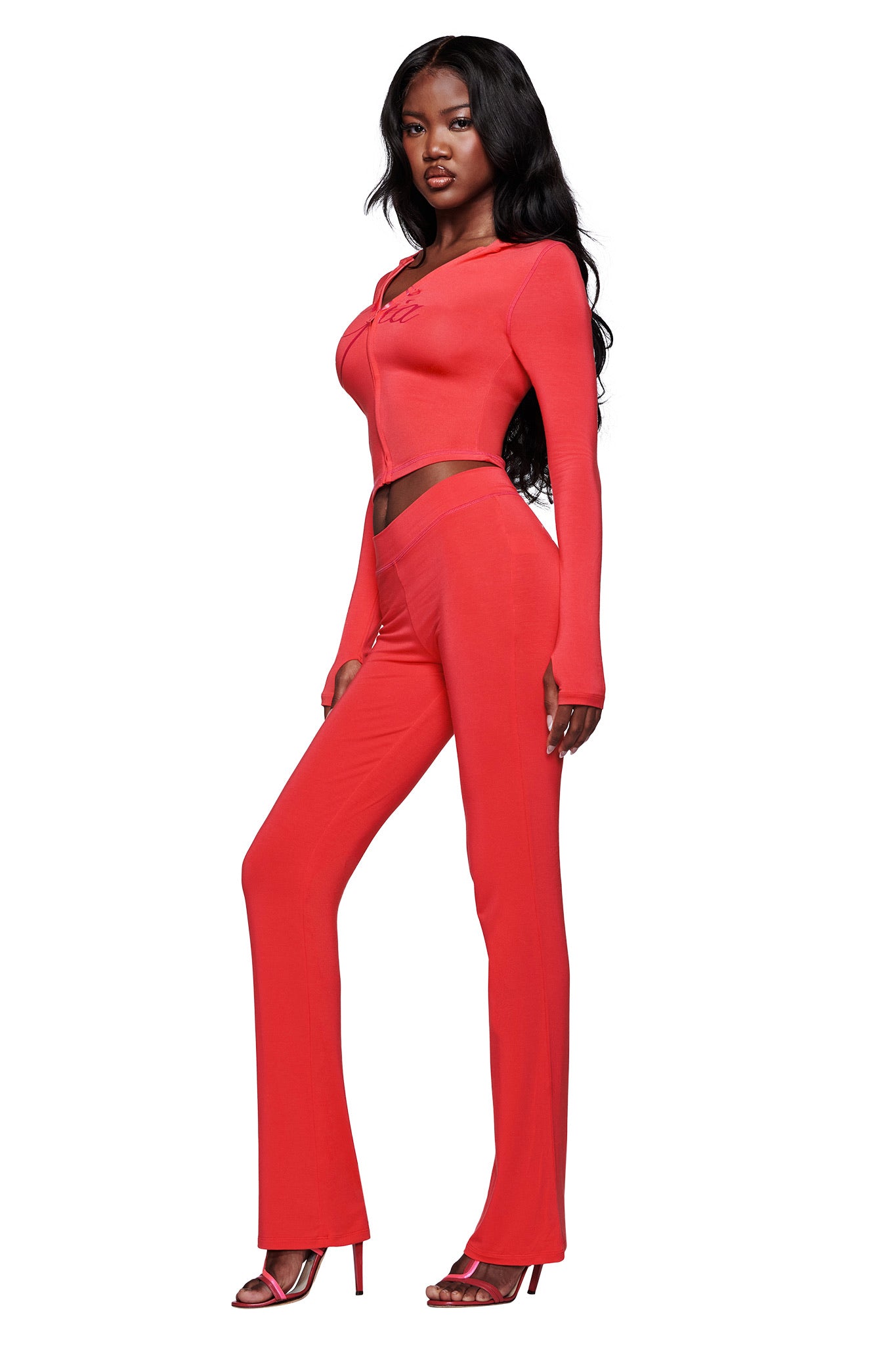 A woman with long black hair poses against a white background wearing a fitted, bright red two-piece set with long sleeves and flared pants, red heels, and the I.AM.GIA BLARE HOODIE in Papaya.