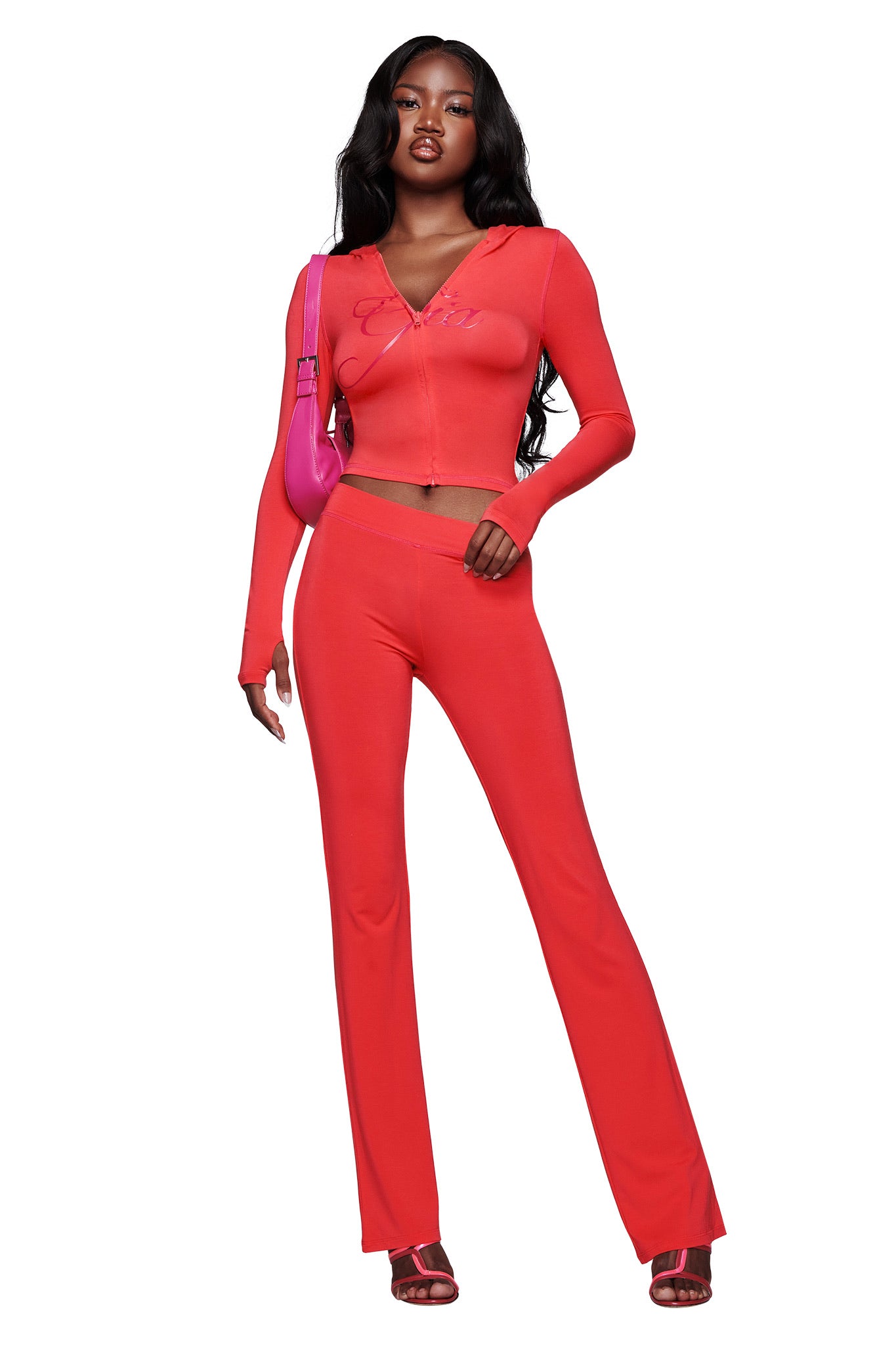 A woman poses against a white background wearing I.AM.GIA's BLARE TRACKPANT in papaya, styled as a bright fitted two-piece set with long sleeves and flared pants, paired with red heeled sandals and a pink shoulder bag.