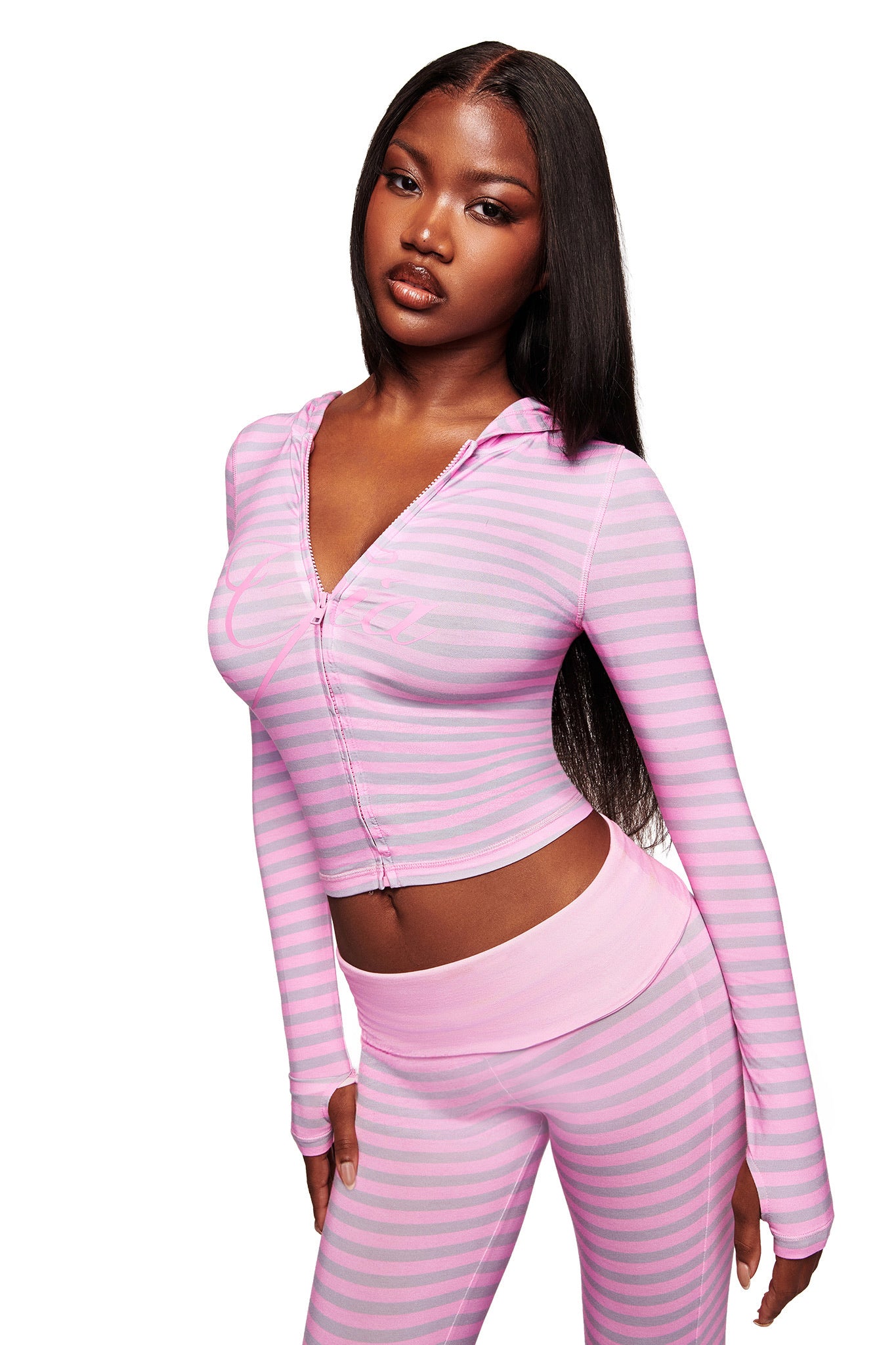 A woman with long straight hair wears the I.AM.GIA BLARE HOODIE in light pink stripe with matching high-waisted leggings, standing against a white background.