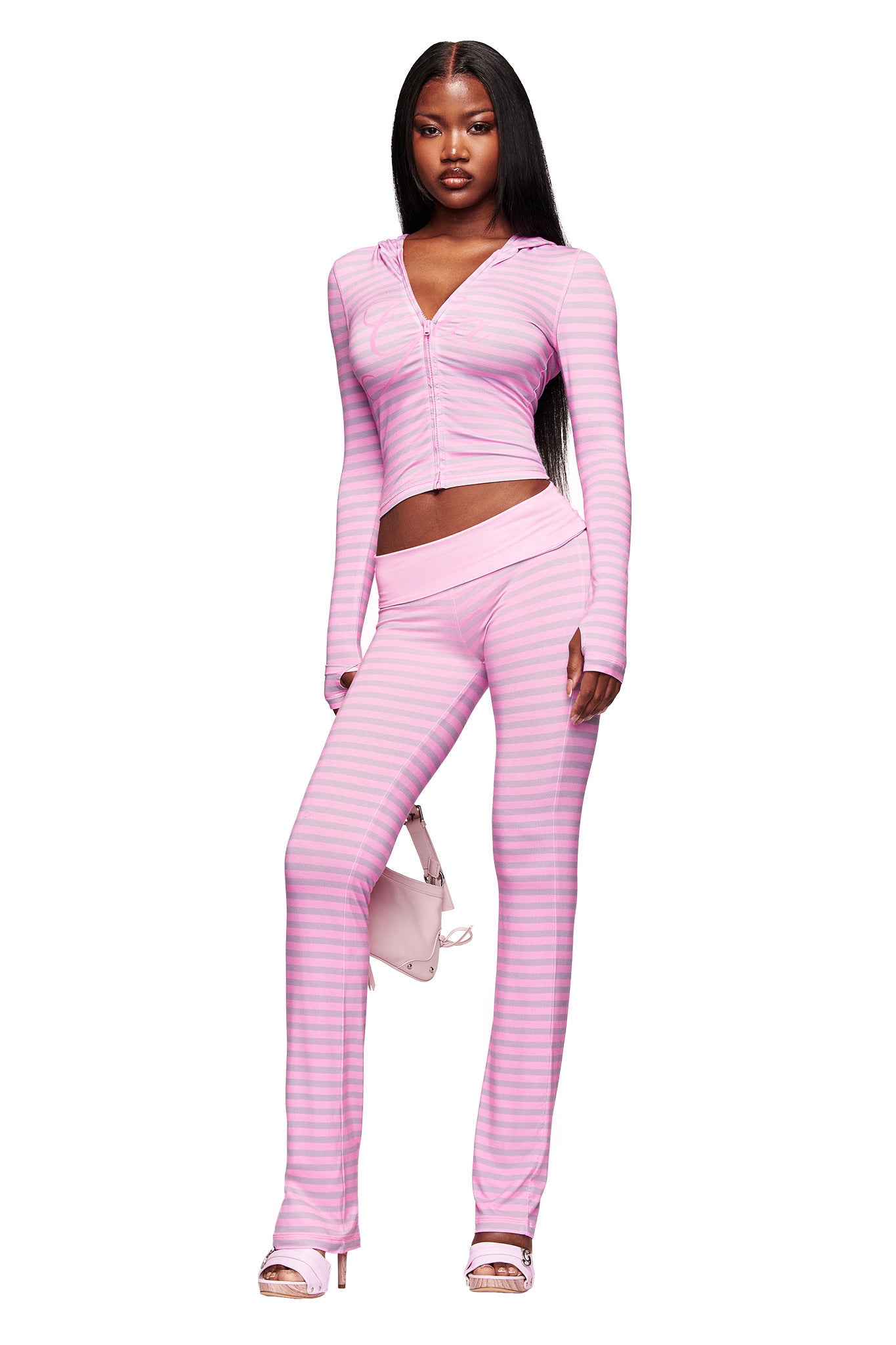 A woman exudes confidence in the I.AM.GIA BLARE FOLDOVER TRACKPANT - LIGHT PINK STRIPE, styled with a fitted, striped two-piece set, a pink purse, and heeled sandals.