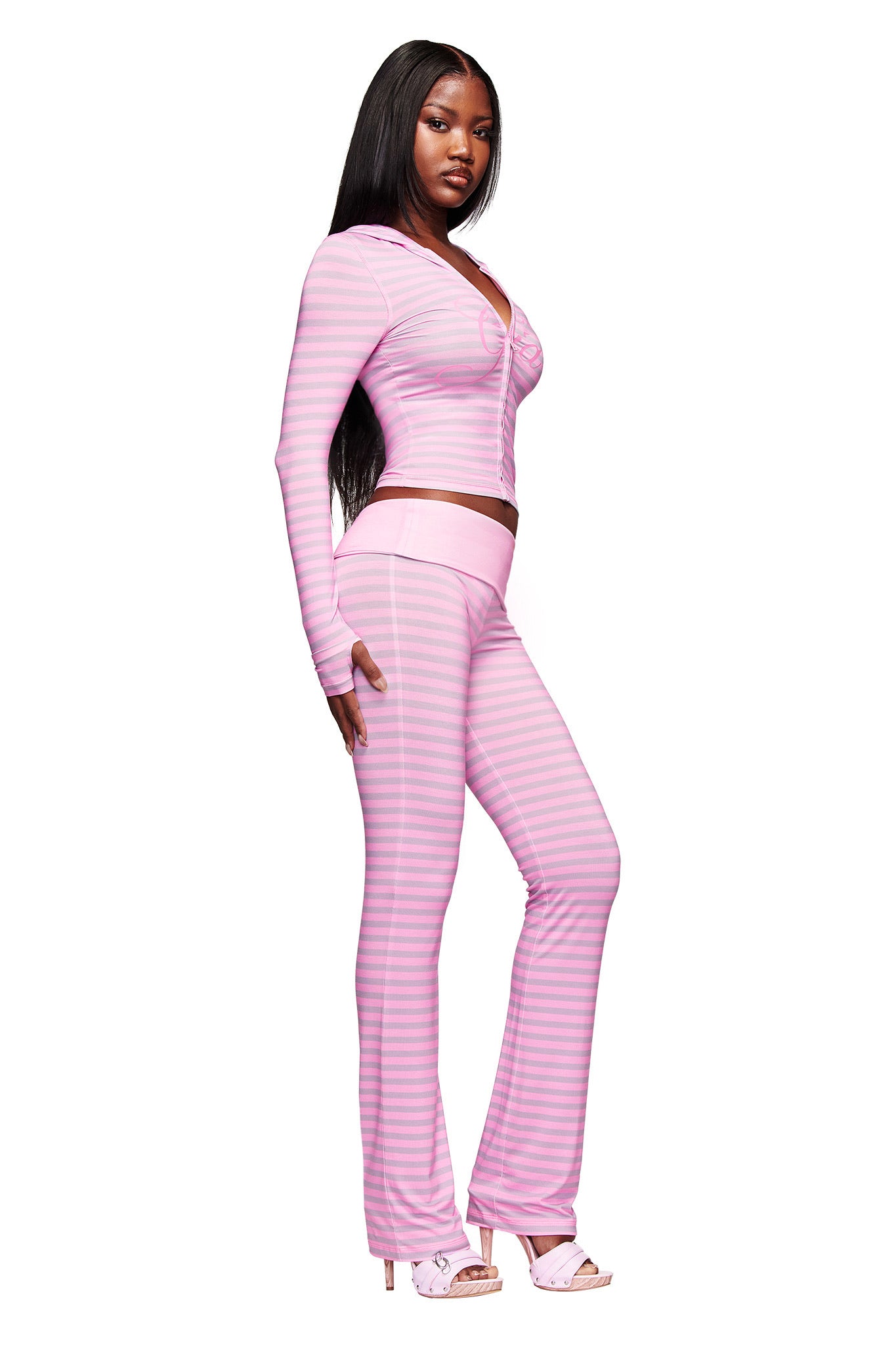A woman stands in a three-quarter pose wearing the I.AM.GIA BLARE FOLDOVER TRACKPANT - LIGHT PINK STRIPE, featuring light and dark pink horizontal stripes, a flared leg, and foldover waistband, paired with pink high-heeled sandals.