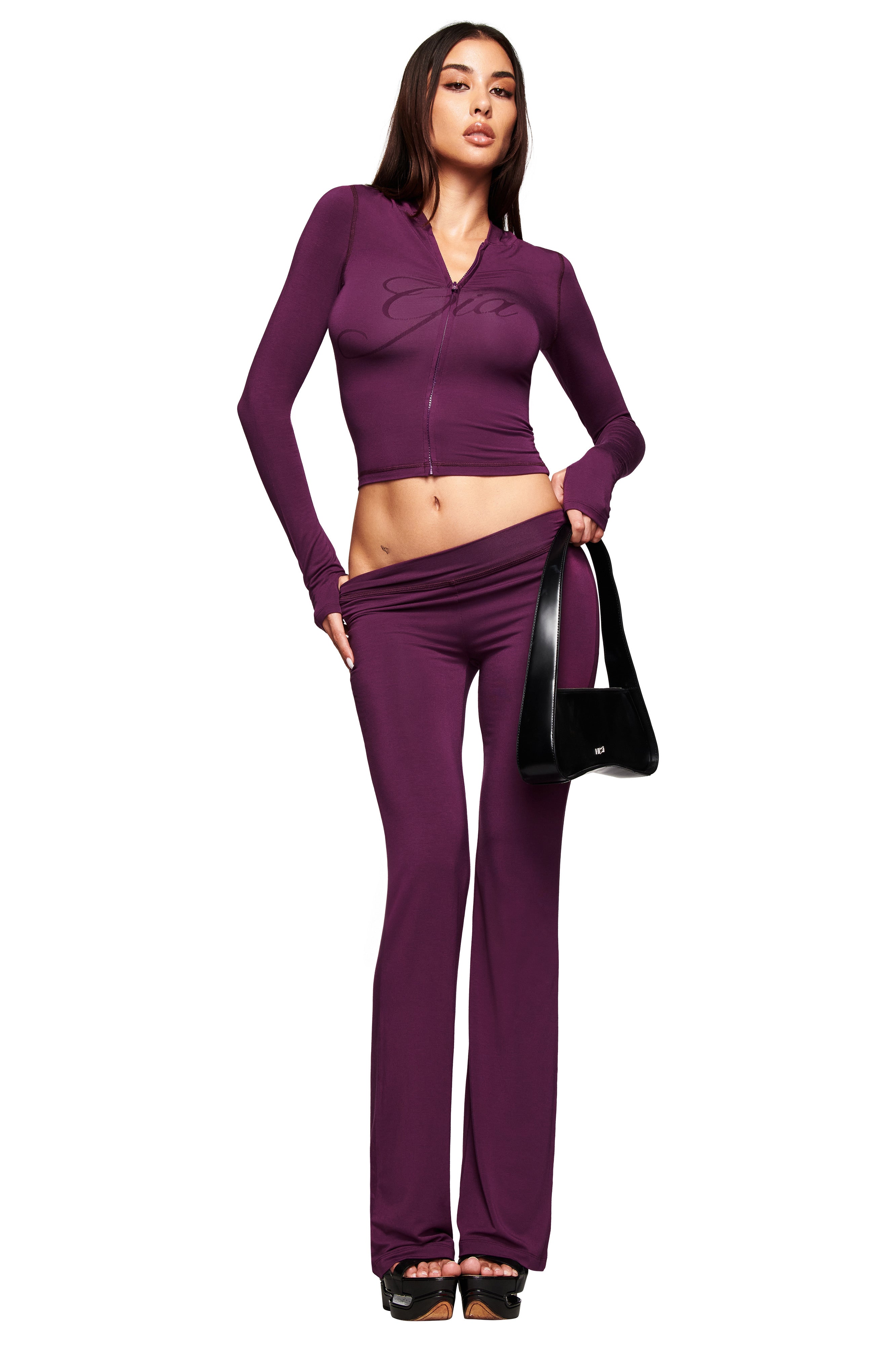 A woman poses in I.AM.GIA's BLARE TRACKPANT - PLUM, a low-rise plum trackpant paired with a fitted, long-sleeve butterfly crop top. She holds a black handbag and lowers the waistband to showcase her hip.