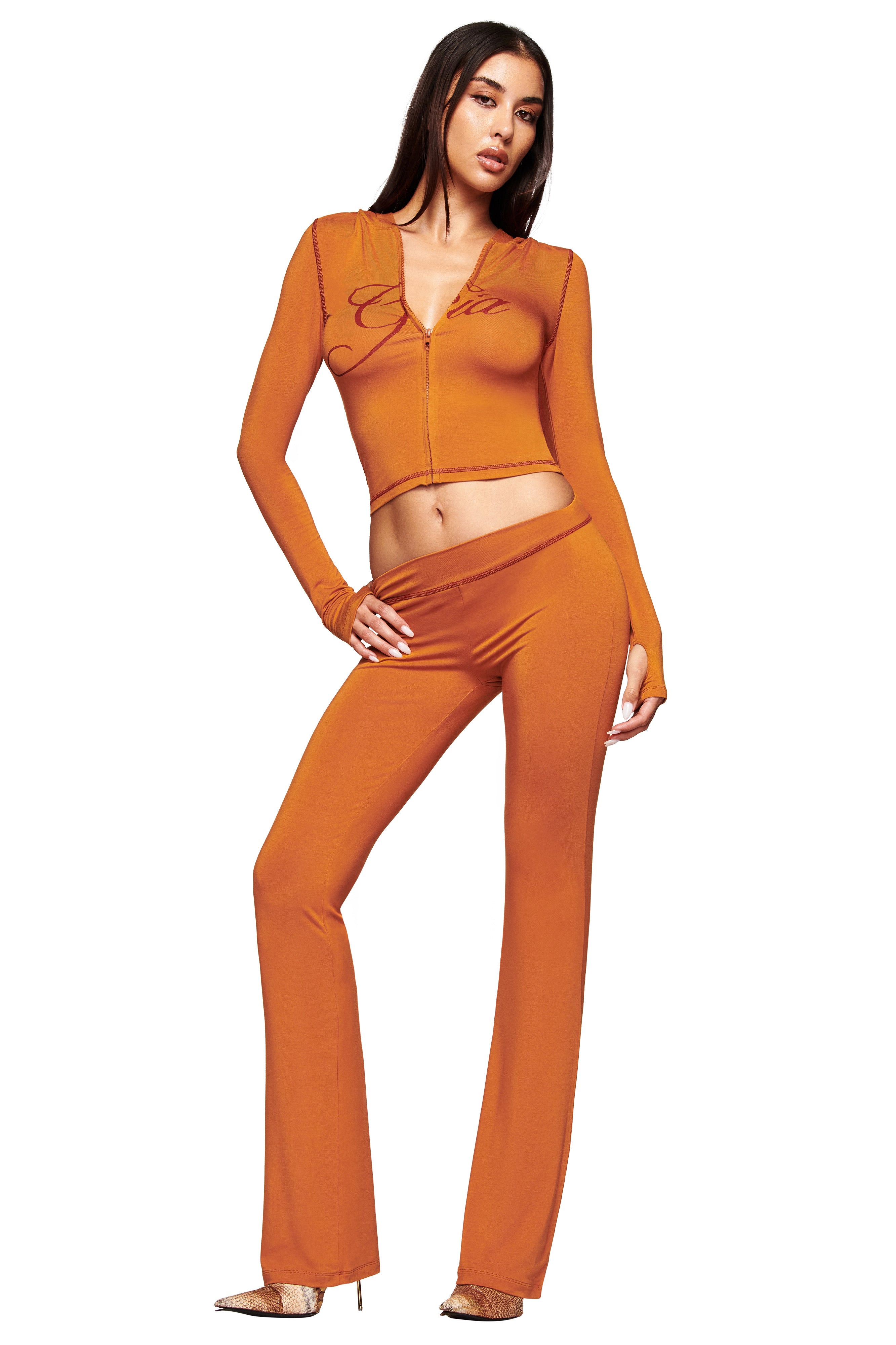A woman poses confidently in the I.AM.GIA BLARE TRACKPANT - PUMPKIN and a matching orange zip-front fitted crop top, both with a bodycon silhouette, paired with heels against a plain white background.