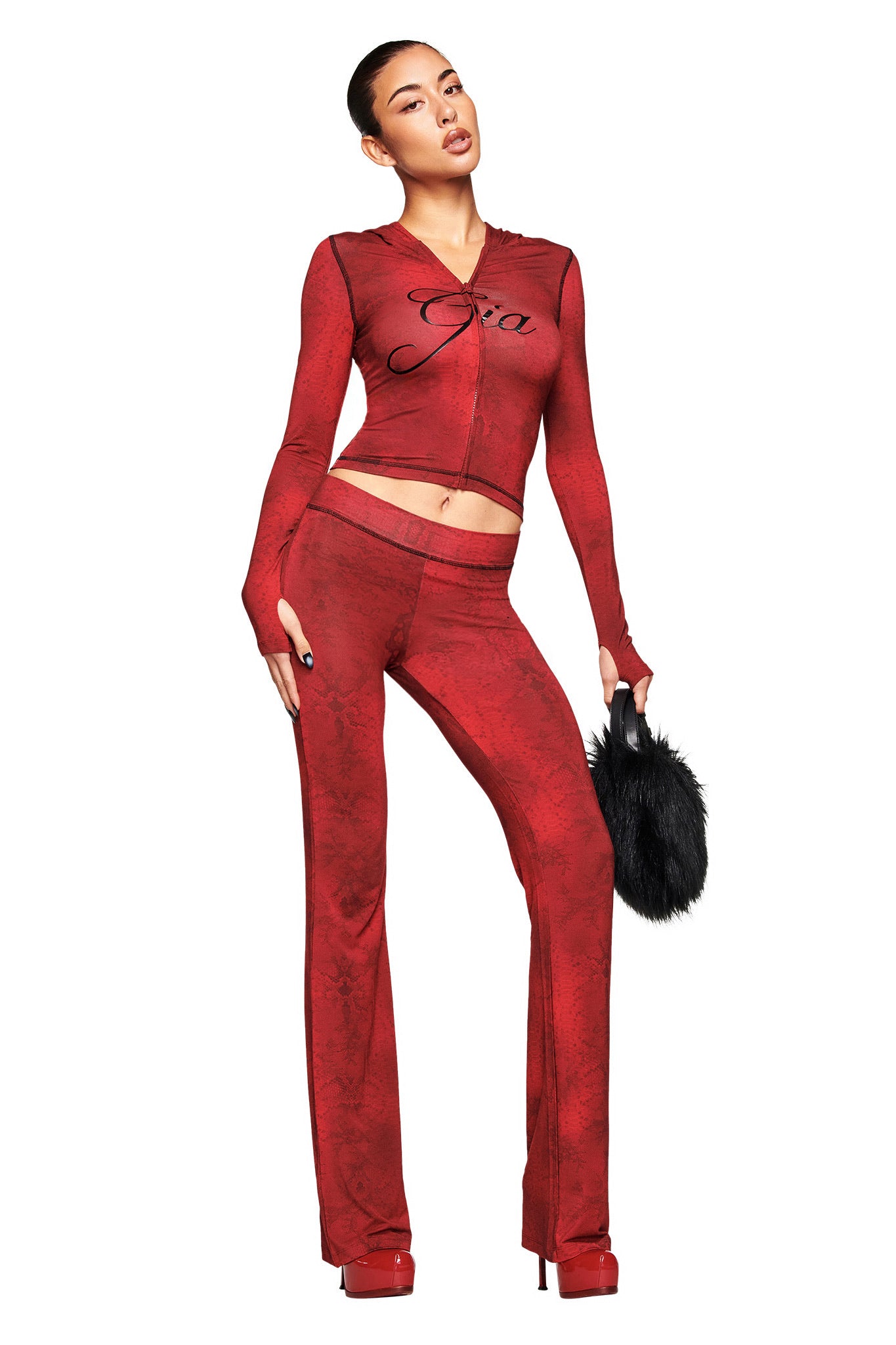 A woman models the I.AM.GIA BLARE HOODIE in red serpentine print, paired with matching flared pants and red platform heels. She holds a black furry handbag and stands against a white background with her hair pulled back.