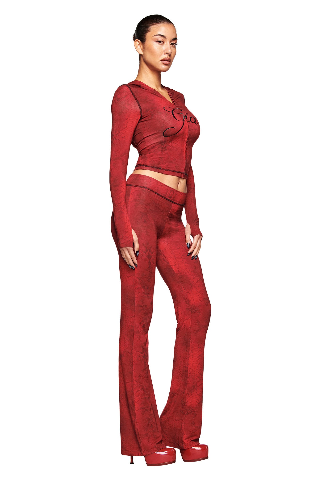 A woman models the BLARE HOODIE - RED SERPENTINE by I.AM.GIA, a fitted, long-sleeve crop top with red serpentine print, paired with matching flared pants and red platform heels against a crisp white background.