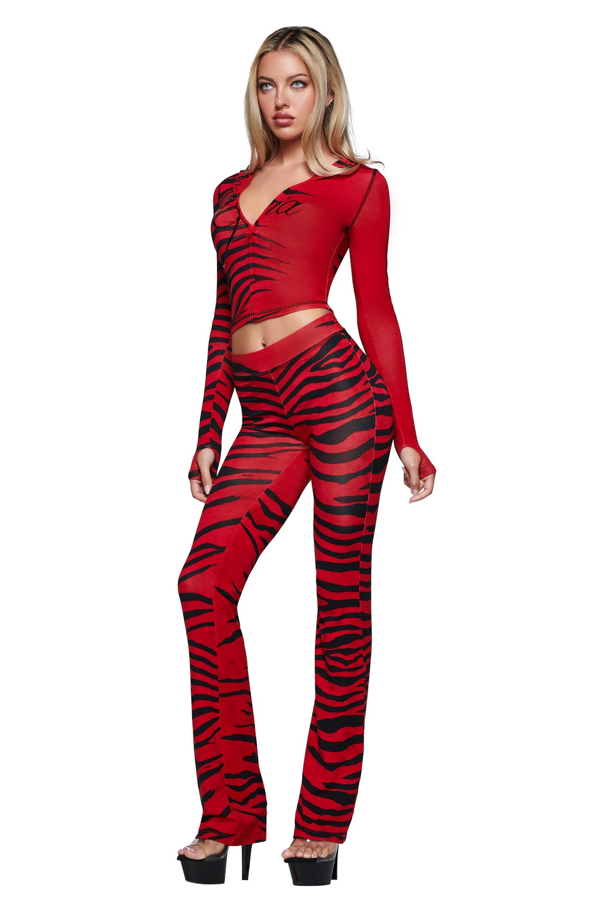 A woman stands against a white background wearing the I.AM.GIA BLARE TRACKPANT - RED WILD SIDE, featuring bold black tiger stripes and the brand’s logo for a striking statement look. She pairs the fitted, long-sleeve red top and matching trackpants with black high-heeled shoes.