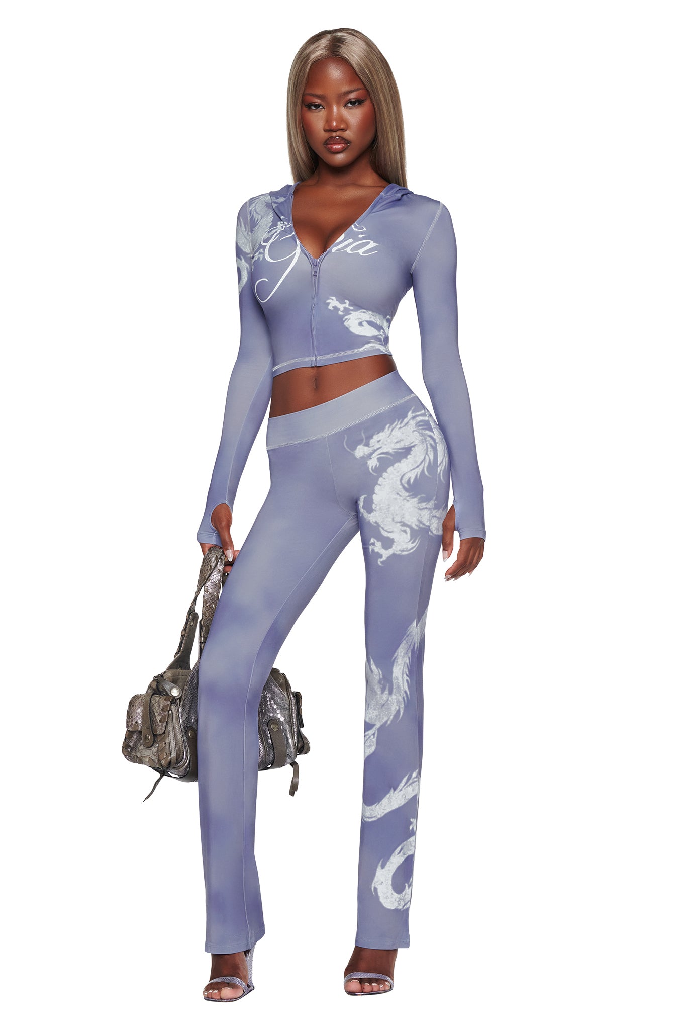 A woman stands confidently in the I.AM.GIA BLARE HOODIE - STEEL BLUE DRAGON, a fitted light purple two-piece featuring white steel blue dragon designs, accessorized with a textured handbag and heeled sandals.