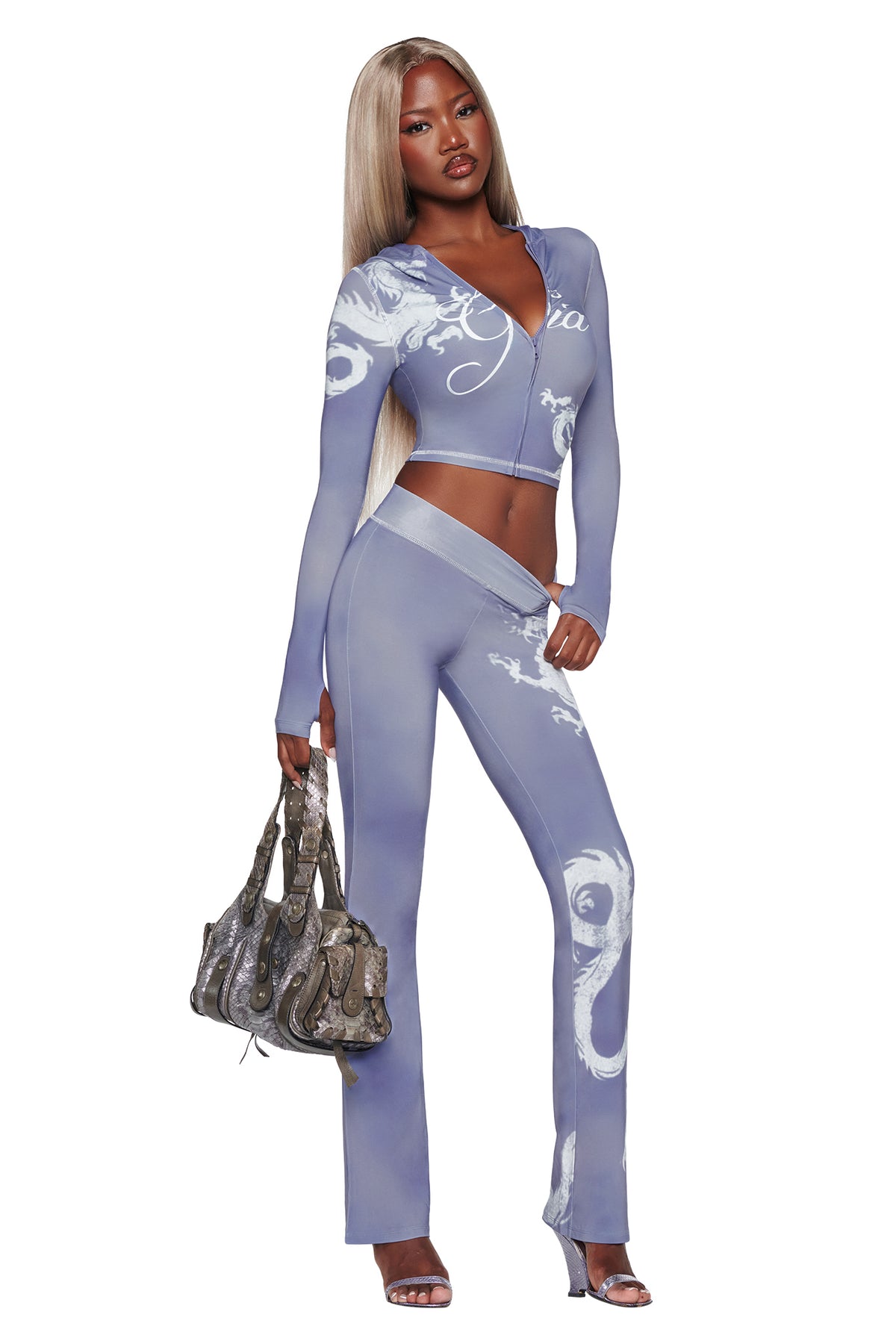 Wearing the I.AM.GIA BLARE HOODIE in Steel Blue Dragon, a woman with long blonde hair poses confidently against a white background. She completes her look with silver heels and carries a textured handbag, highlighting the hoodie’s distinctive white dragon-inspired pattern on its light purple, fitted two-piece outfit.