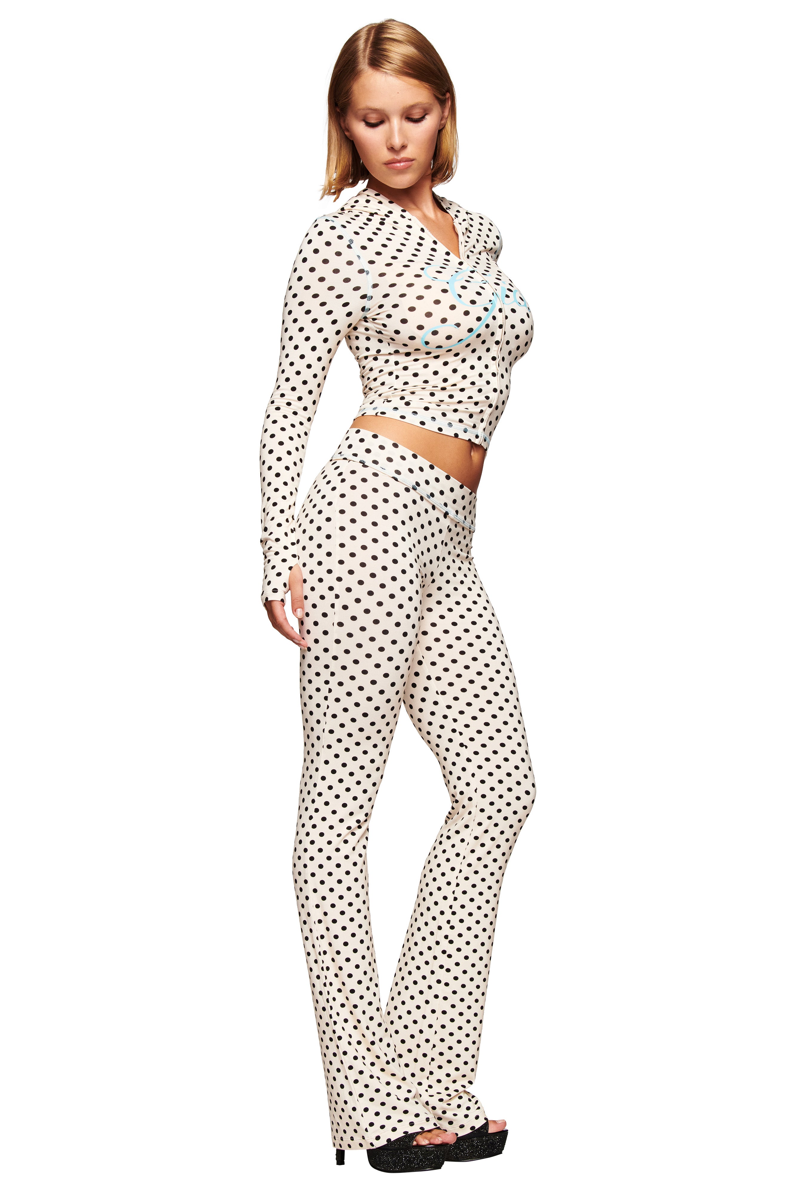 A woman poses in the I.AM.GIA BLARE TRACKPANT - CREAM POLKA DOT and a matching fitted, long-sleeve crop top, both featuring black polka dots on white. She wears black platform shoes. The pants have flared legs. The background is white.