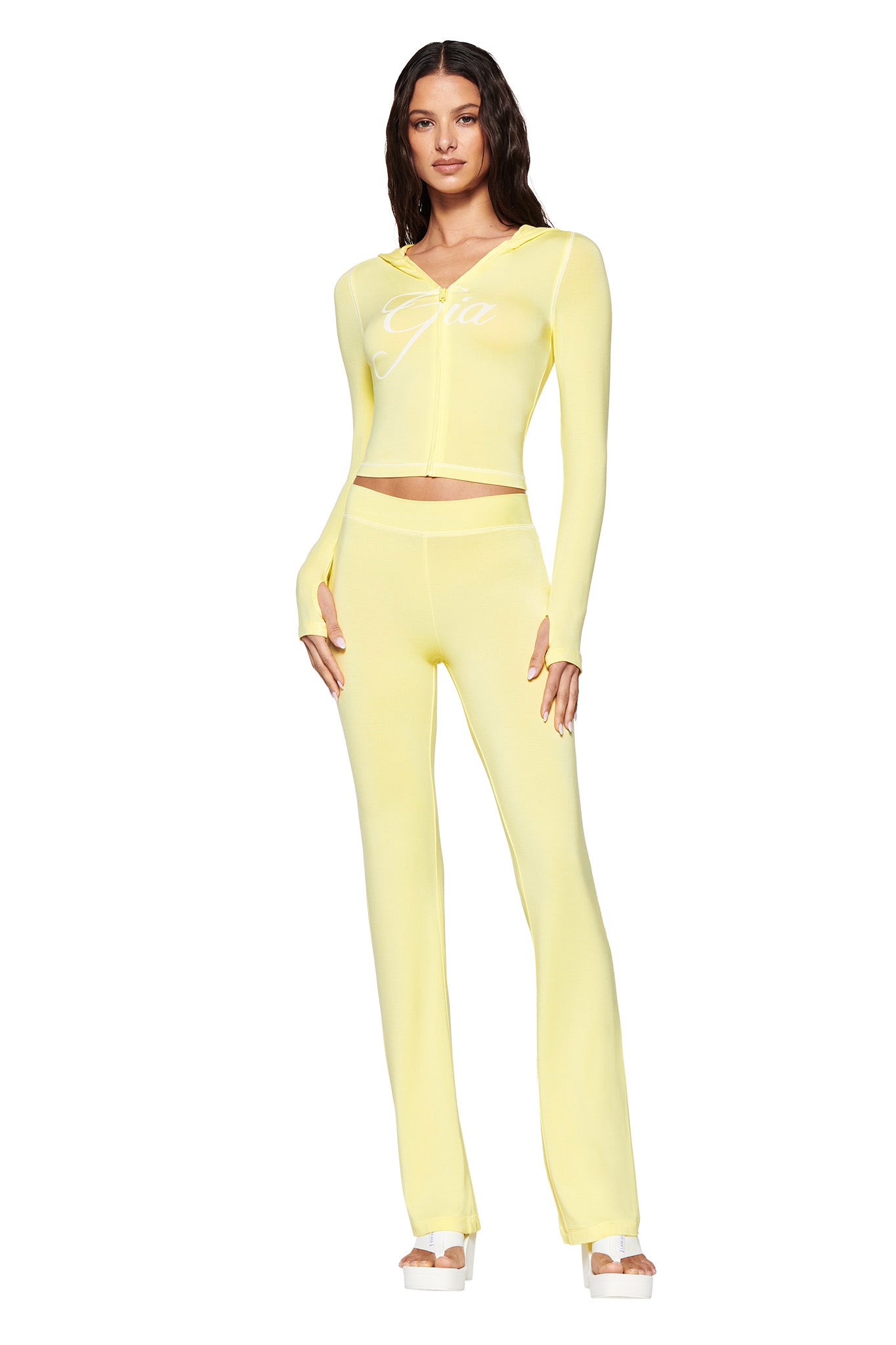 A woman stands gracefully, wearing a matching pale yellow outfit from I.AM.GIA featuring a long-sleeve top with "Gia" and fitted BLARE TRACKPANT. Her arms rest by her sides, highlighting the bodycon silhouette, while her long hair flows down. She wears sandals against the crisp white background.
