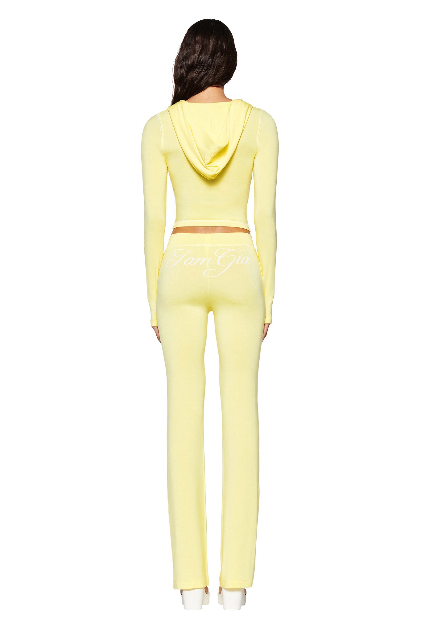 A person with long dark hair wears the I.AM.GIA Blare Hoodie and matching pants in pale yellow, featuring a thumb-hole design. The outfit is perfectly paired with white platform shoes, set against a crisp white background.