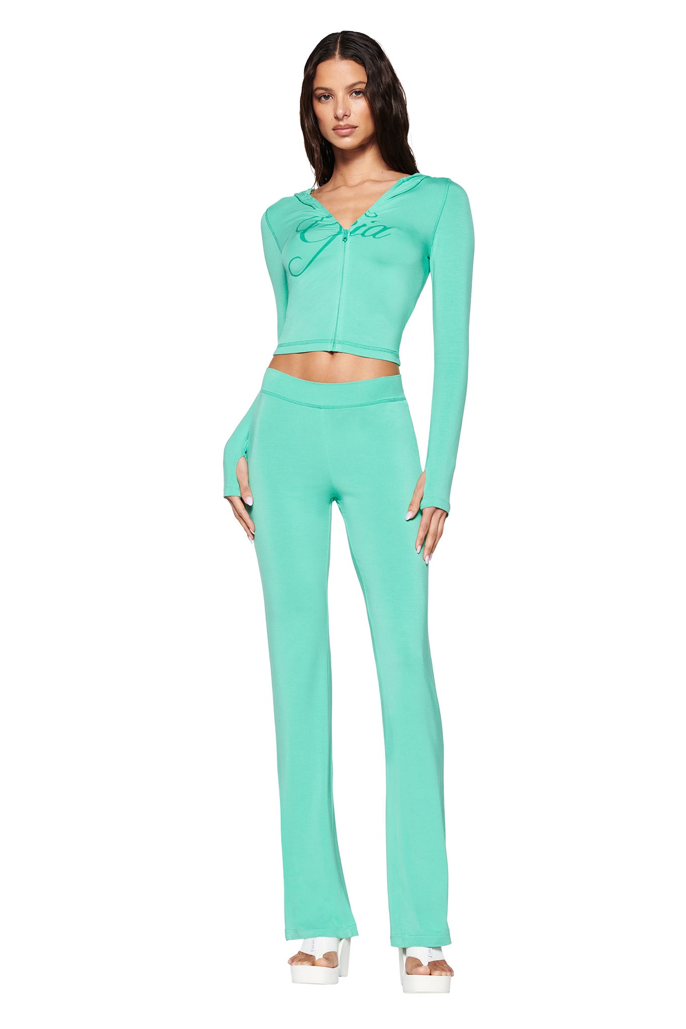 A woman in a teal zip-up long-sleeve top with the I.AM.GIA logo and matching BLARE TRACKPANT - GREEN : TURQUOISE : TEAL stands in platform sandals against a white background.