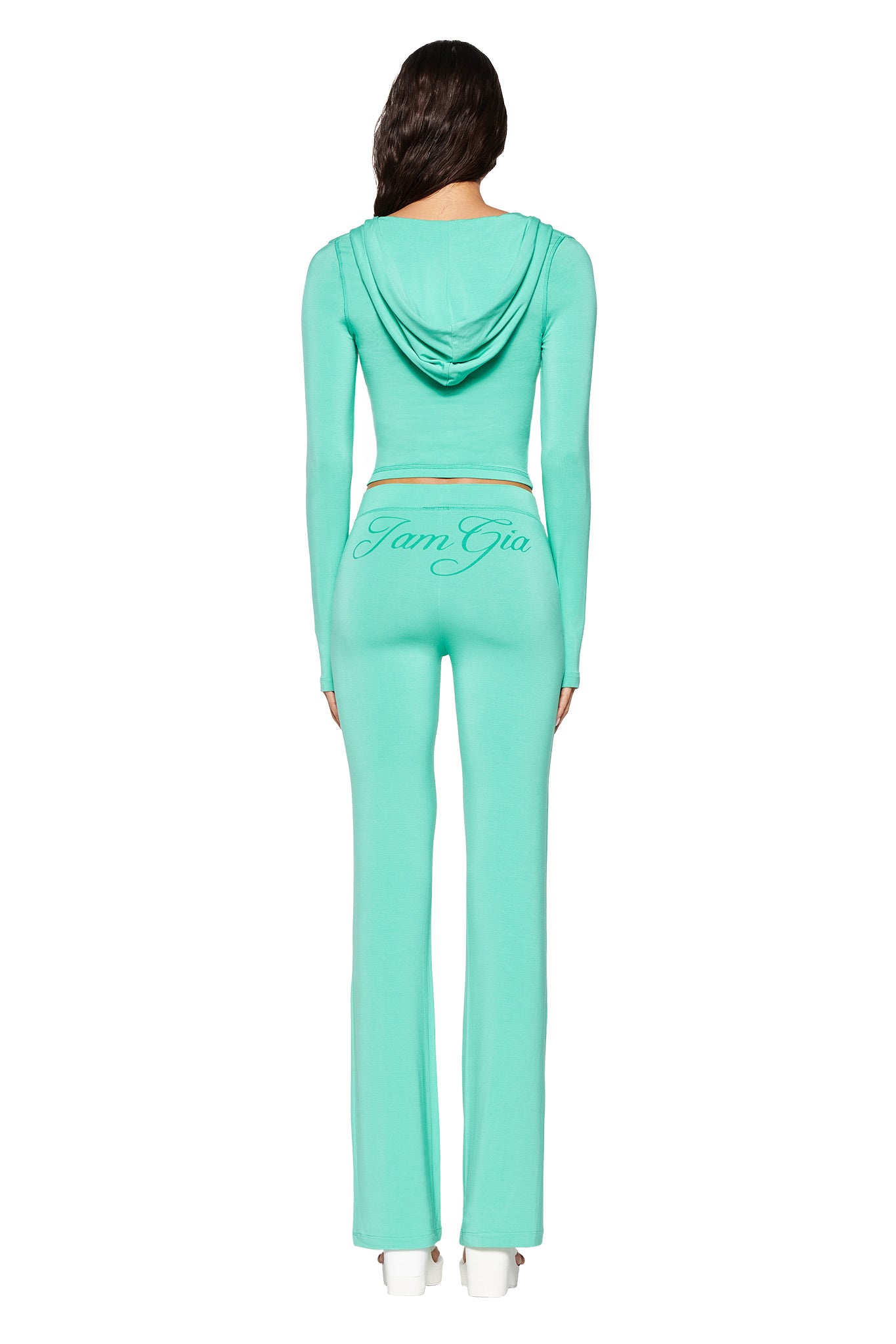A person with long hair, seen from behind, wears a light turquoise Blare hoodie and matching pants from I.AM.GIA. The bodycon fit highlights the casual yet fitted style, and "I am Gia" is printed on the back of the pants.