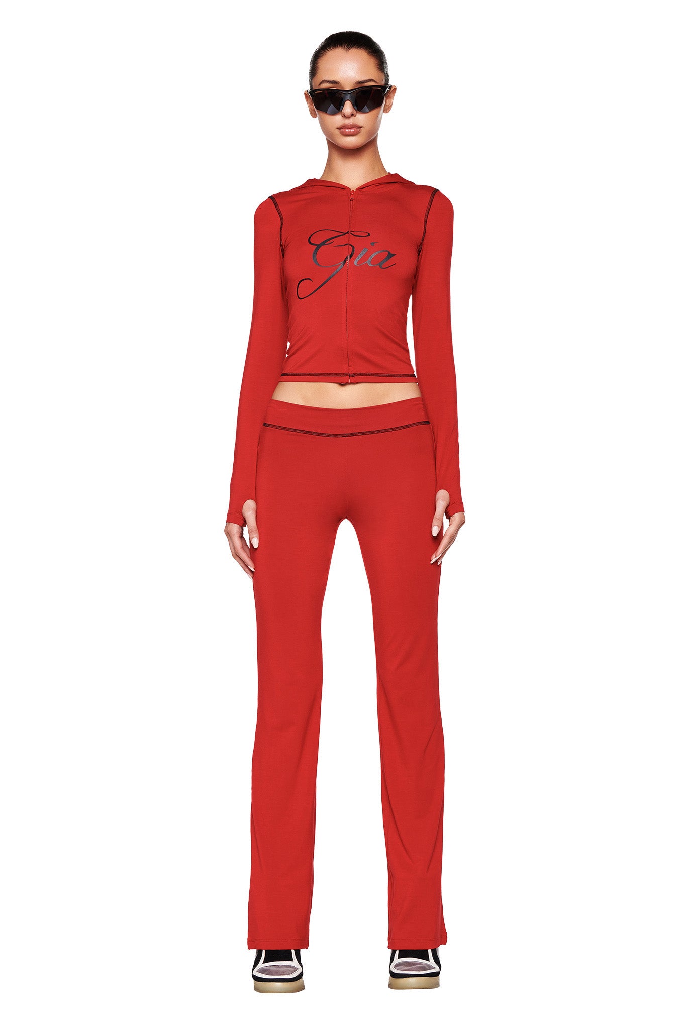 A person stands facing forward wearing a fitted red zip-up hoodie made from stretchy bamboo fabric and matching I.AM.GIA BLARE TRACKPANT - RED. The hoodie features text across the chest area. The person is also dressed in black sunglasses and black-and-white sneakers, with their hands placed at their sides.