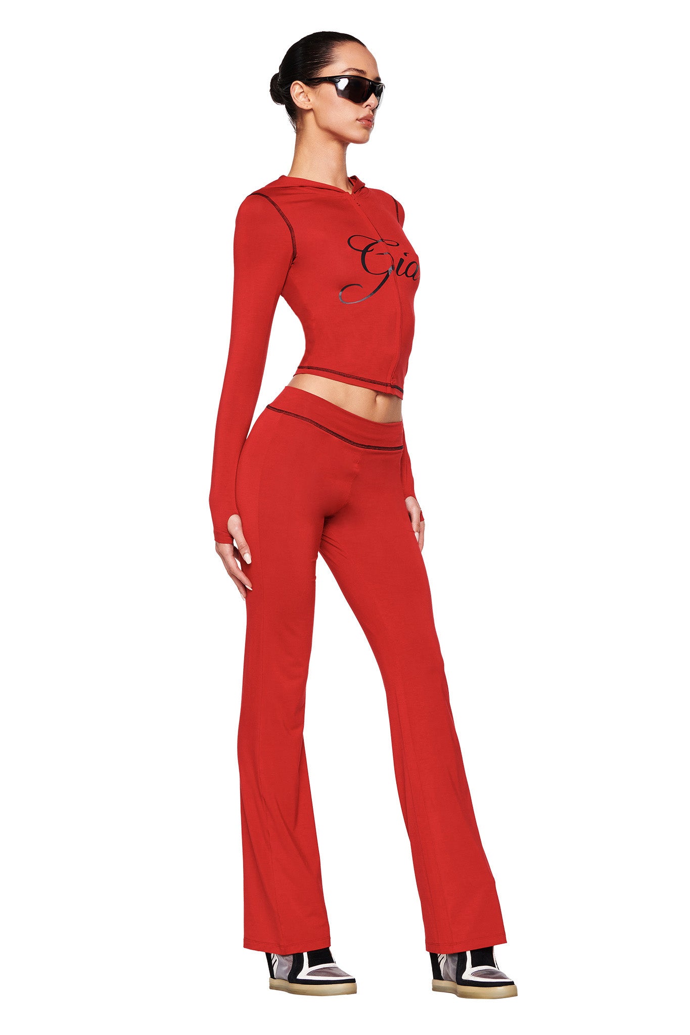A person with dark hair tied back is wearing a vibrant red outfit from I.AM.GIA, made of stretchy bamboo fabric. The ensemble includes a long-sleeved crop top with black detailing and matching BLARE TRACKPANTS in red. Additionally, they are accessorized with black sunglasses and high-top sneakers, posing against a white background.