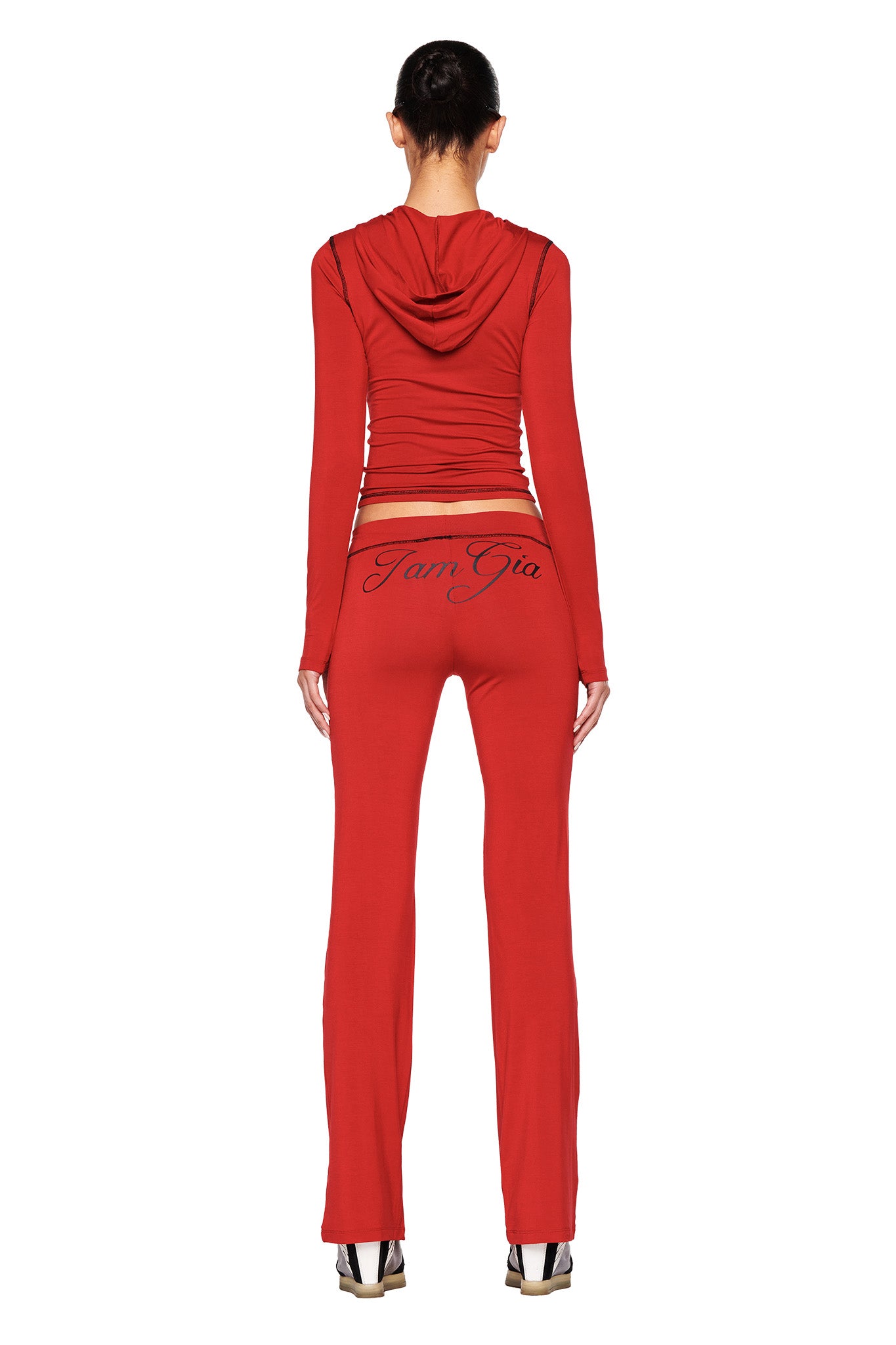 A person is shown from the back, wearing a matching red set of a hooded long-sleeve top and the BLARE TRACKPANT - RED by I.AM.GIA with "Iam Gia" written on the back. The outfit features a bodycon silhouette, paired with high-heeled shoes that have a rectangular, white-patterned design.