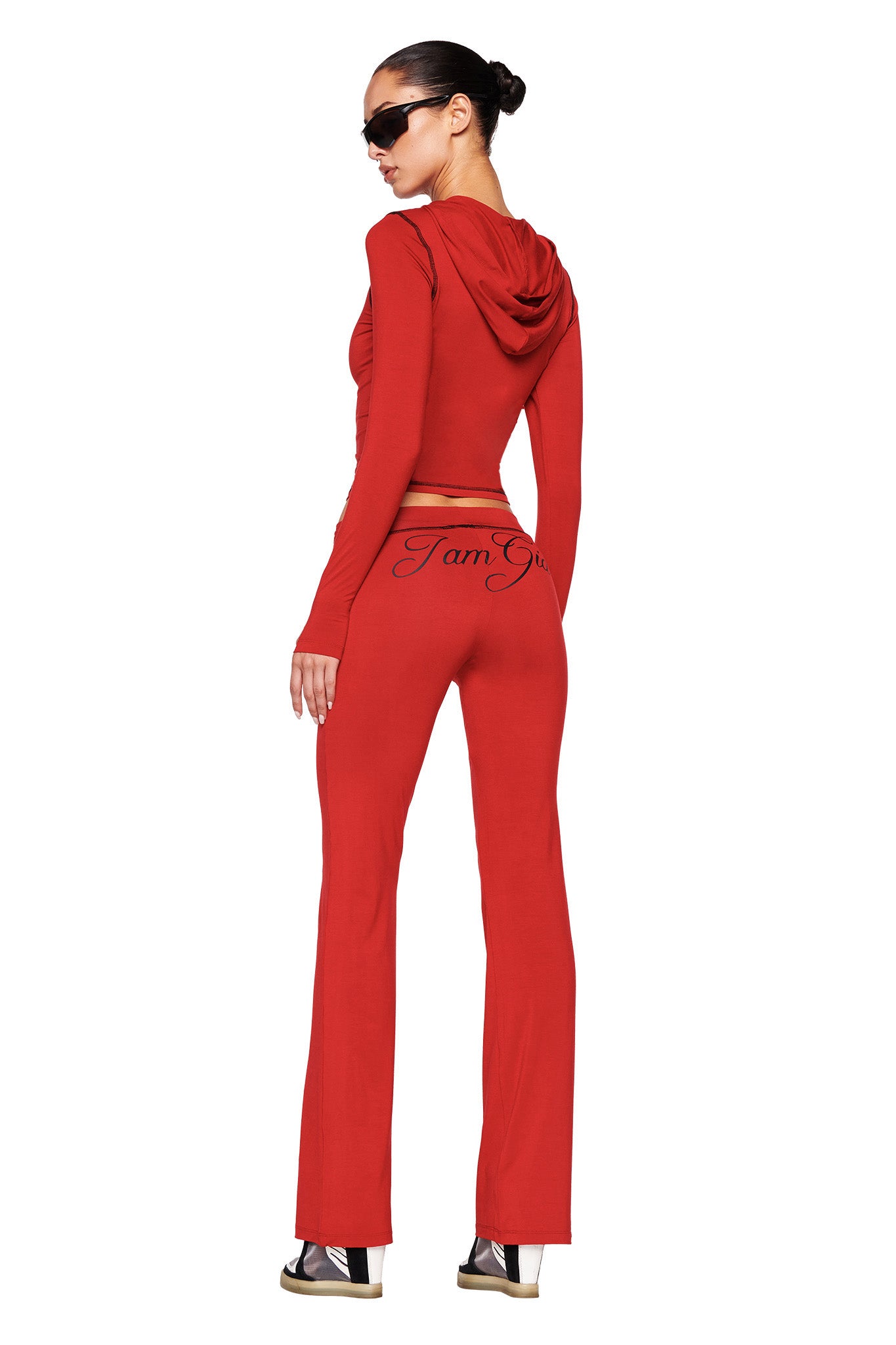 A person is facing away, sporting the red Blare Hoodie crop top by I.AM.GIA paired with matching high-waisted pants featuring "Team Girl" printed on the back. They complement their outfit with black platform sandals and dark sunglasses, their hair styled in a sleek bun. The bodycon silhouette emphasizes their figure perfectly.