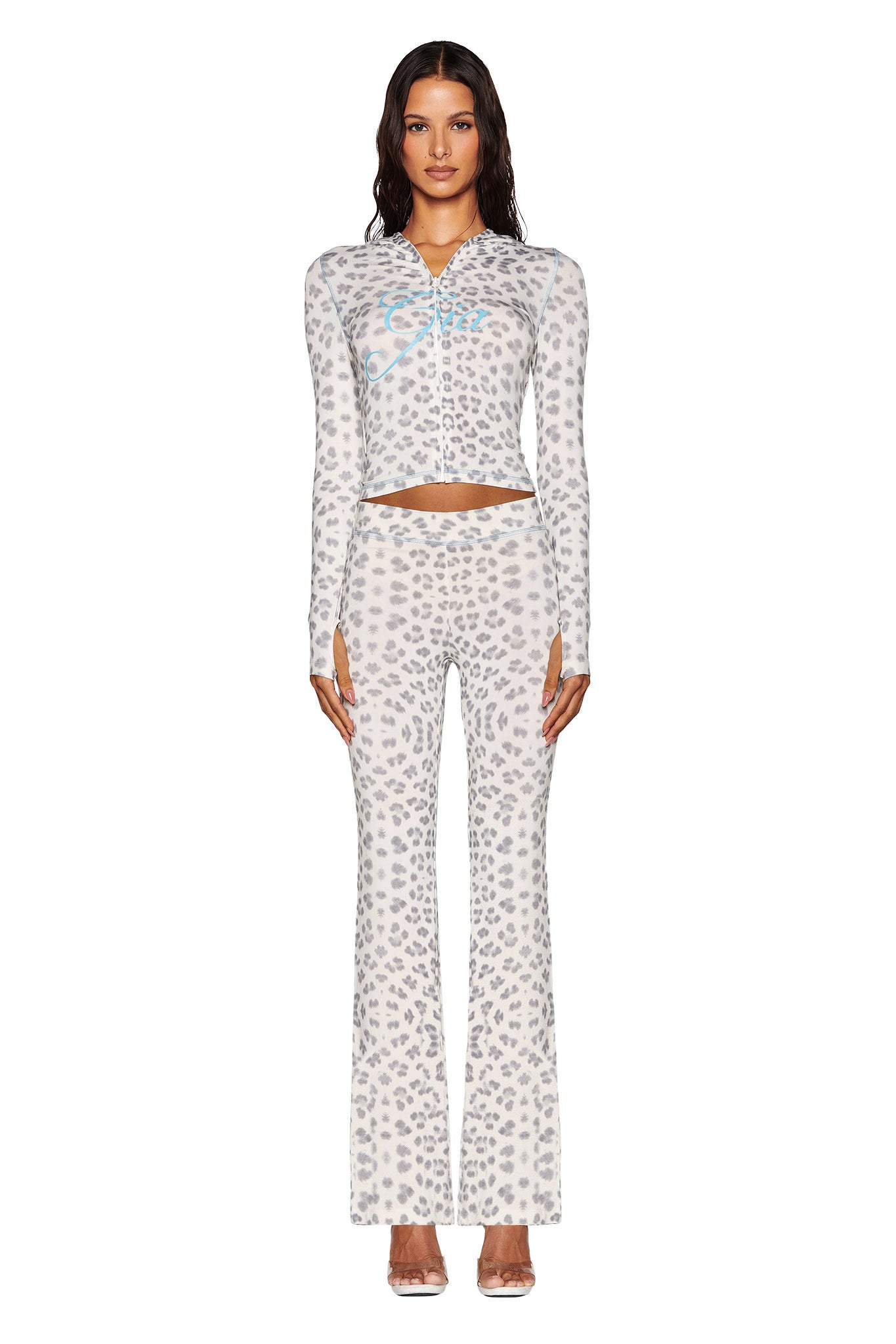 A person wearing the I.AM.GIA BLARE HOODIE - SNOW LEOPARD from the Winter Wild Card collection is seen in a coordinating two-piece outfit. This ensemble includes a bodycon fit long-sleeve top with a front zipper and high-waisted, flared pants, styled against a plain white background.