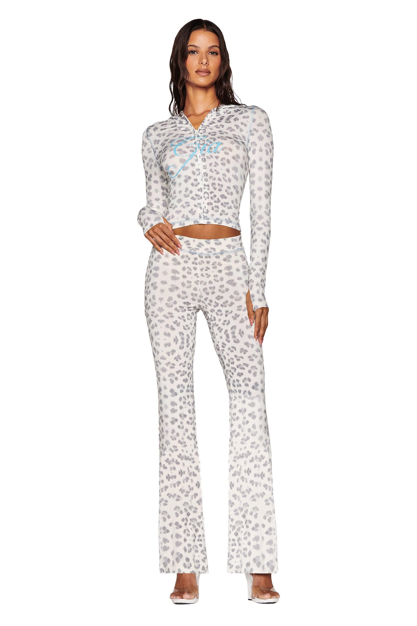 A person confidently stands in the BLARE HOODIE - SNOW LEOPARD from I.AM.GIA's WINTER WILD CARD collection, styled with matching flared pants. The chic ensemble is complemented by clear high-heeled shoes against a plain white background.