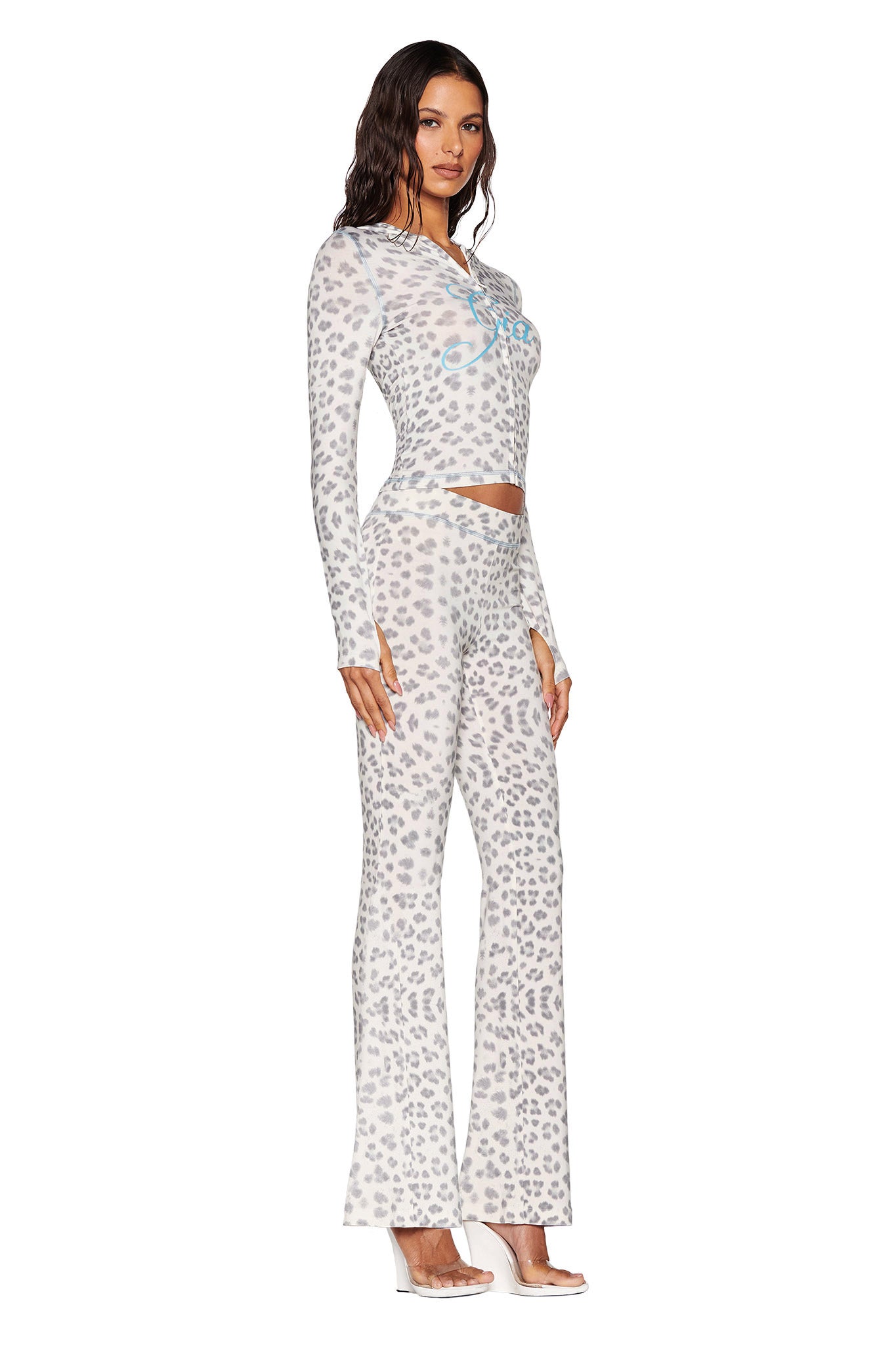 A woman showcases the BLARE HOODIE in a snow leopard design from I.AM.GIA's Winter Wild Card collection. She pairs it with clear high-heeled shoes, standing in profile with her hair styled in loose waves.