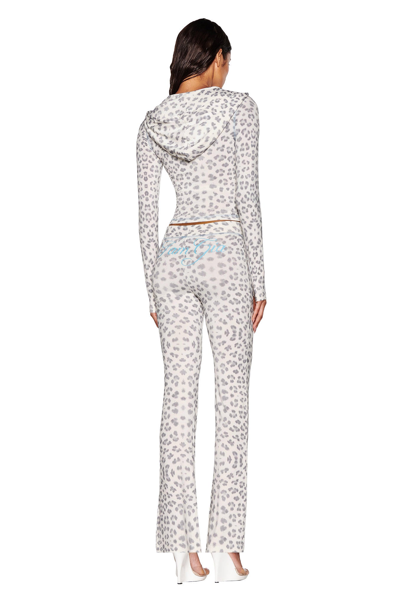 A person is seen from behind wearing a snow leopard print outfit, consisting of a hooded top and the BLARE TRACKPANT - SNOW LEOPARD : WINTER WILD CARD, paired with white high heels. Against a plain white background, the I.AM.GIA logo is subtly showcased on their ensemble.