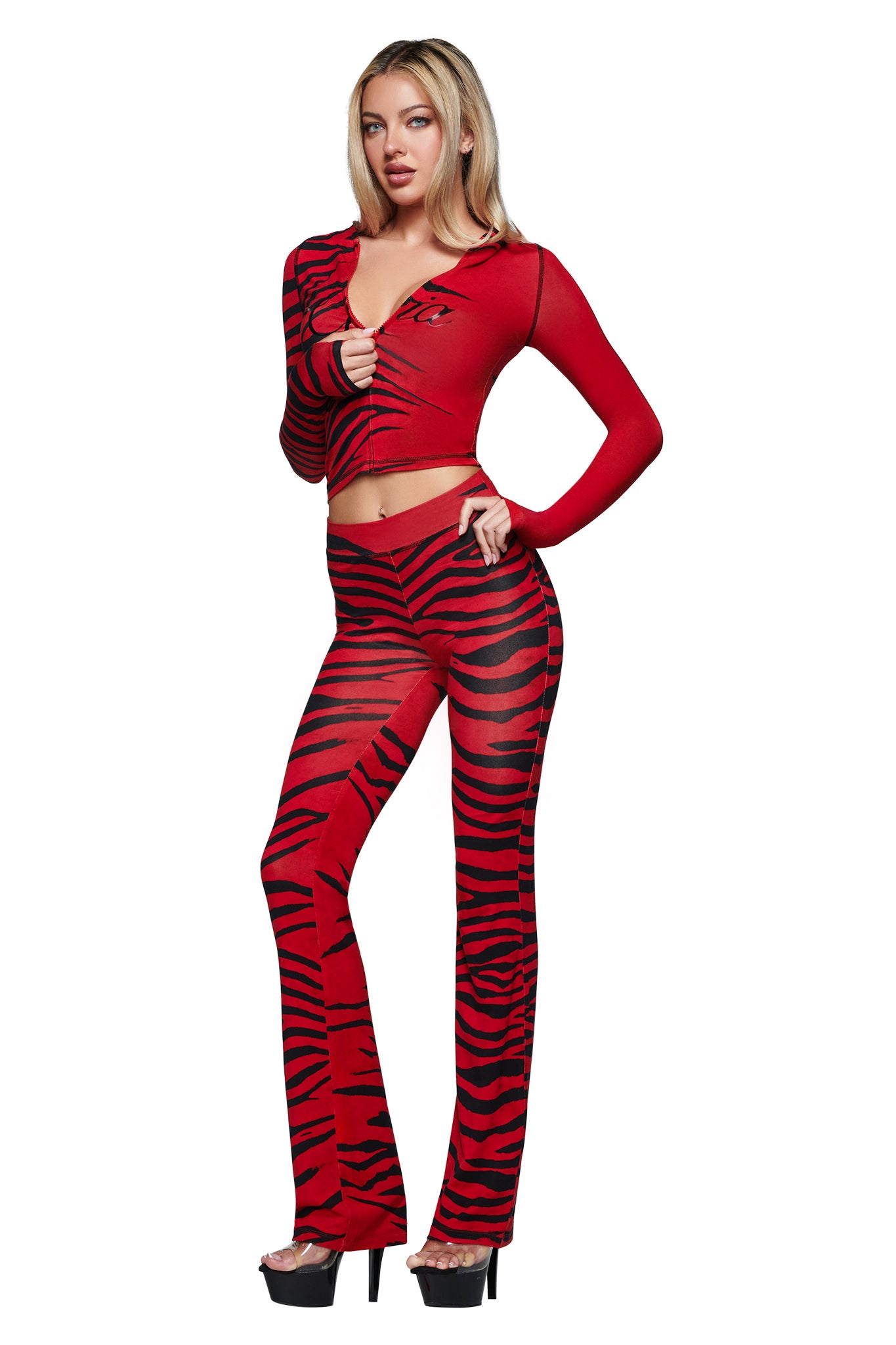 A person wearing the BLARE TRACKPANT - RED WILD SIDE by I.AM.GIA, featuring bold red zebra print and a streetwear-inspired silhouette, styled with a sheer black and red tiger-striped long-sleeved top and flared pants. The look is finished with high heels against a white background.