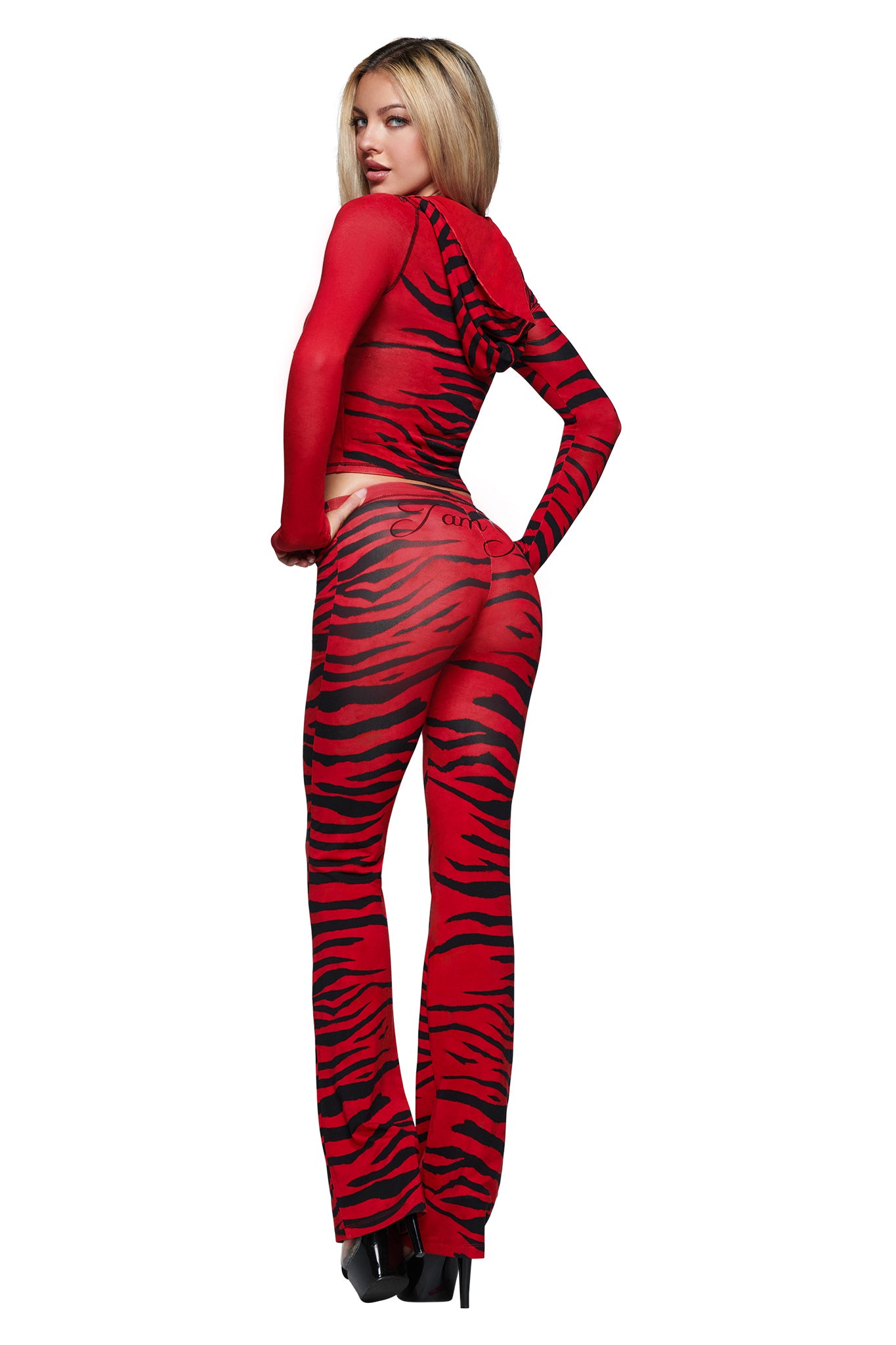 A model with long blonde hair stands in platform heels, wearing the I.AM.GIA BLARE HOODIE in the eye-catching Red Wild Side print—a sheer red and black tiger-striped design. Posing with their back turned and looking over their shoulder, they showcase the bold style of this statement piece.