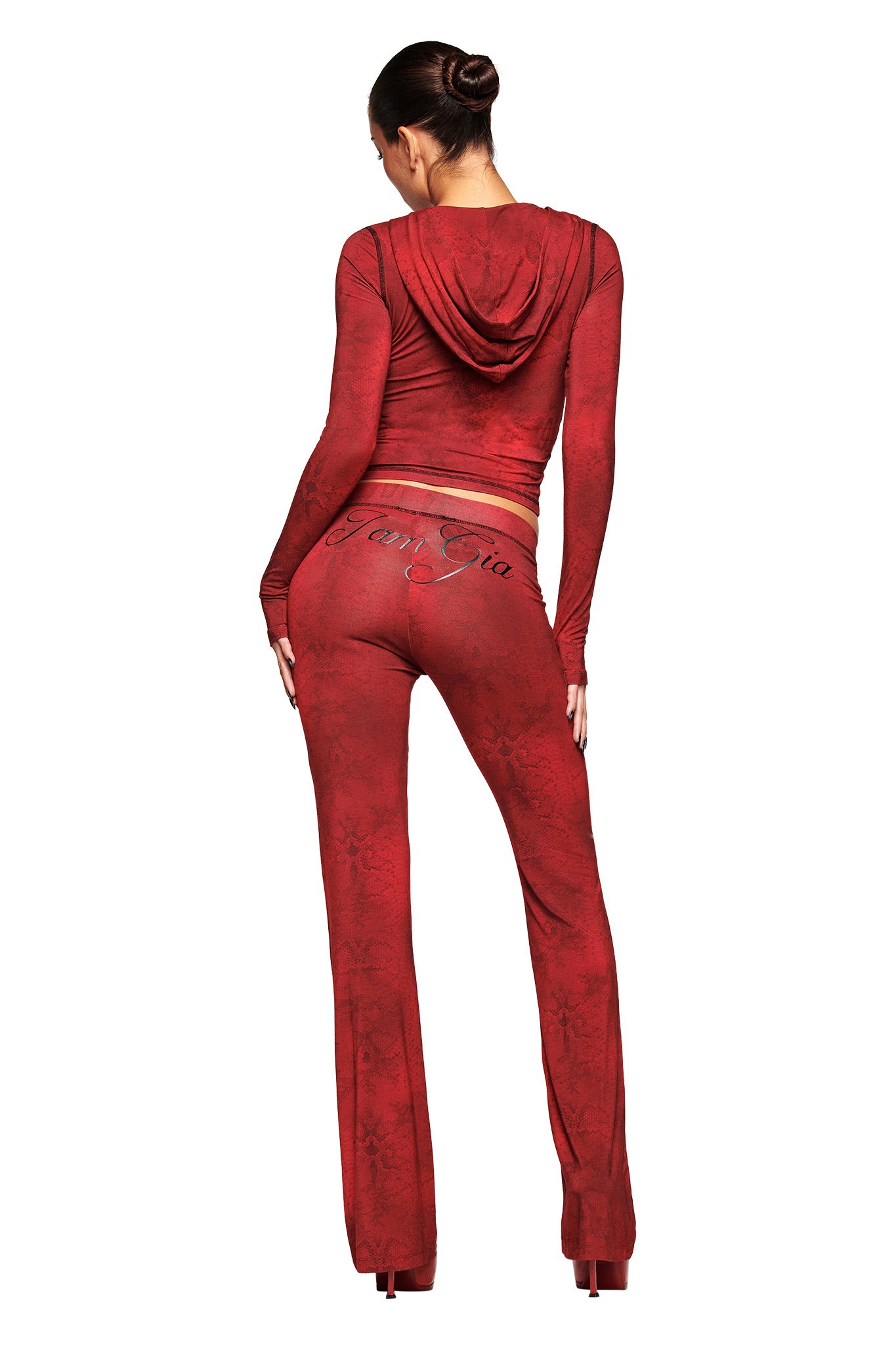 A woman with dark hair in a bun poses with her back turned, wearing I.AM.GIA’s BLARE TRACKPANT - RED SERPENTINE and a matching red hooded top, featuring a low-rise waist and cursive writing on the back against a white background.