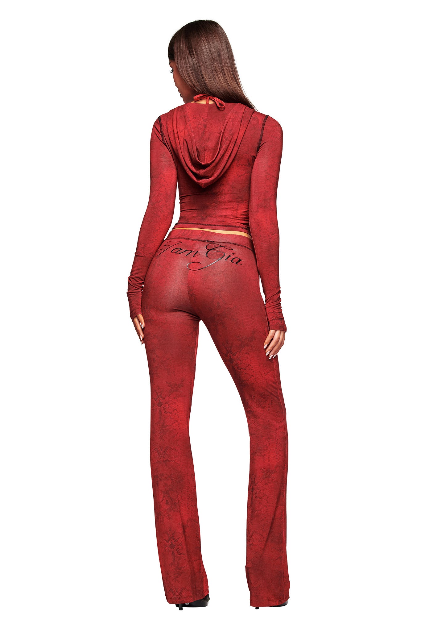 A woman with long brown hair wears the red serpentine BLARE TRACKPANT by I.AM.GIA, featuring "I Am Gia" across the back, paired with a fitted long-sleeve hooded top. She stands facing away against a white background.
