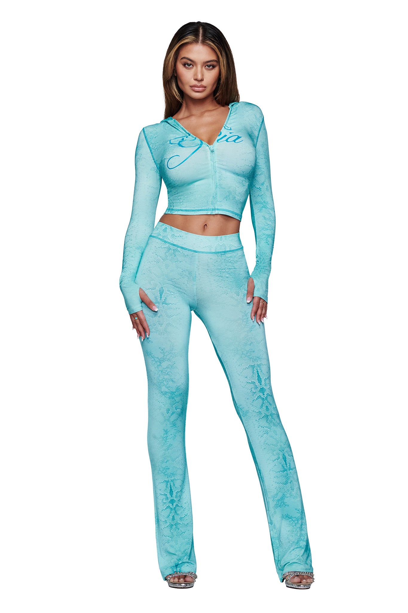 A woman with long, straight hair faces forward wearing the I.AM.GIA BLARE TRACKPANT - TEAL SERPENTINE, styled with a fitted zip-up crop top and silver high-heeled shoes. The pants have a subtle flared leg.