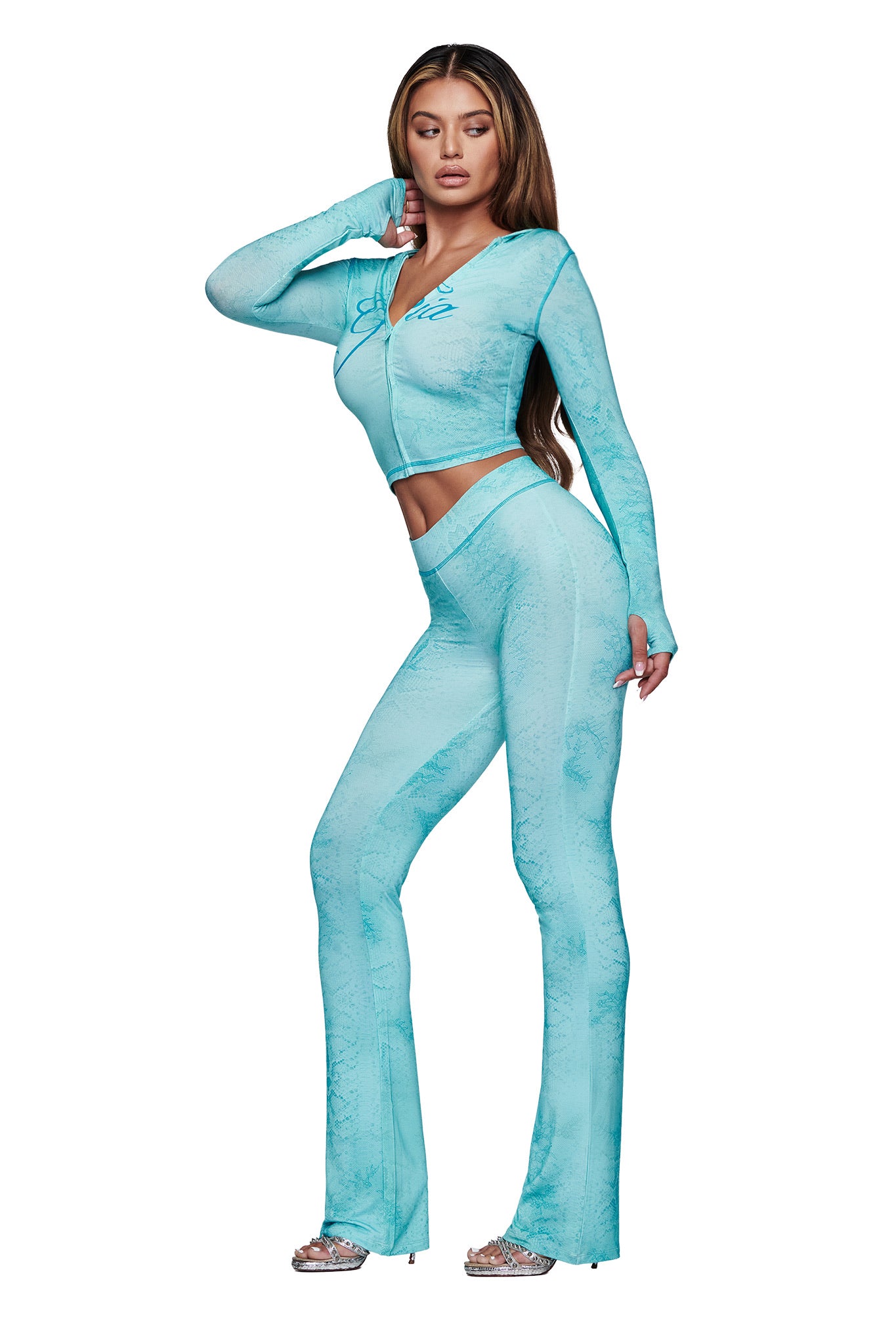 A woman poses in the I.AM.GIA BLARE HOODIE - TEAL SERPENTINE, styled with matching flared pants and silver open-toe heels against a white background.