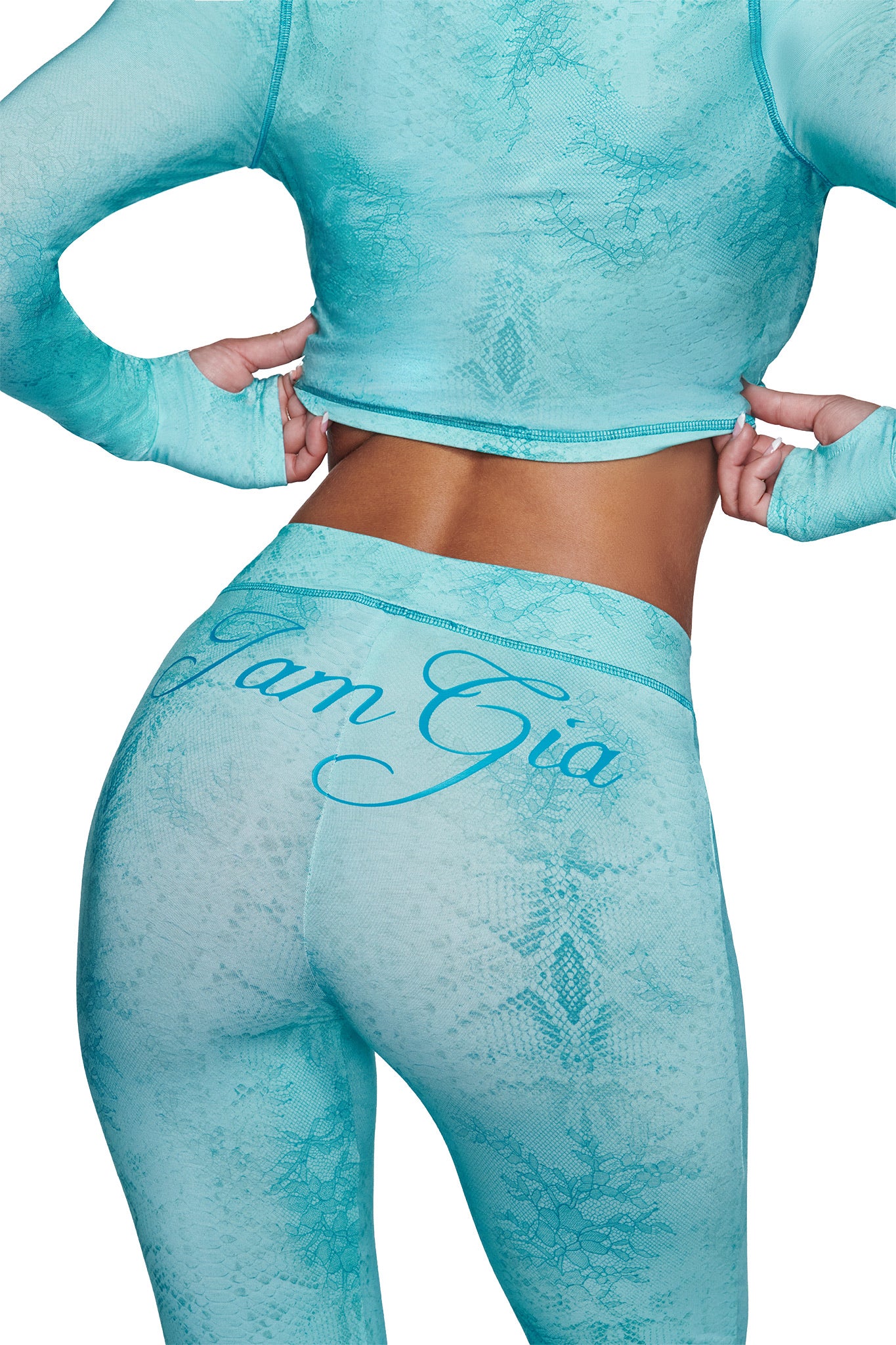 A person is seen from the back wearing a fitted, long-sleeve turquoise top paired with I.AM.GIA's BLARE TRACKPANT - TEAL SERPENTINE leggings, featuring a subtle pattern and "I Am Gia" branding on the waistband.
