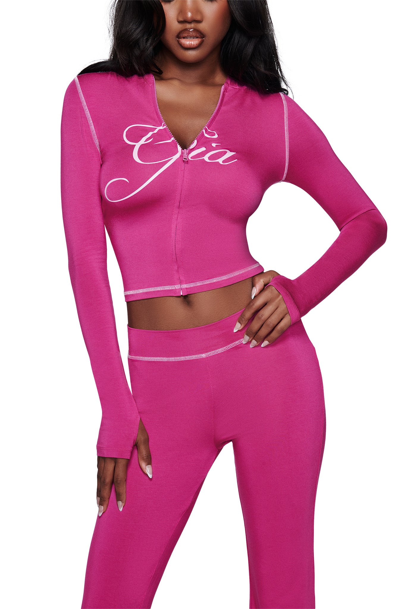 A woman wears the I.AM.GIA BLARE HOODIE in Raspberry, a fitted zip-up top with long sleeves, thumb holes, and white "Gia" script on the chest. Her face is partially out of frame. Matching high-waisted pants complete the look.
