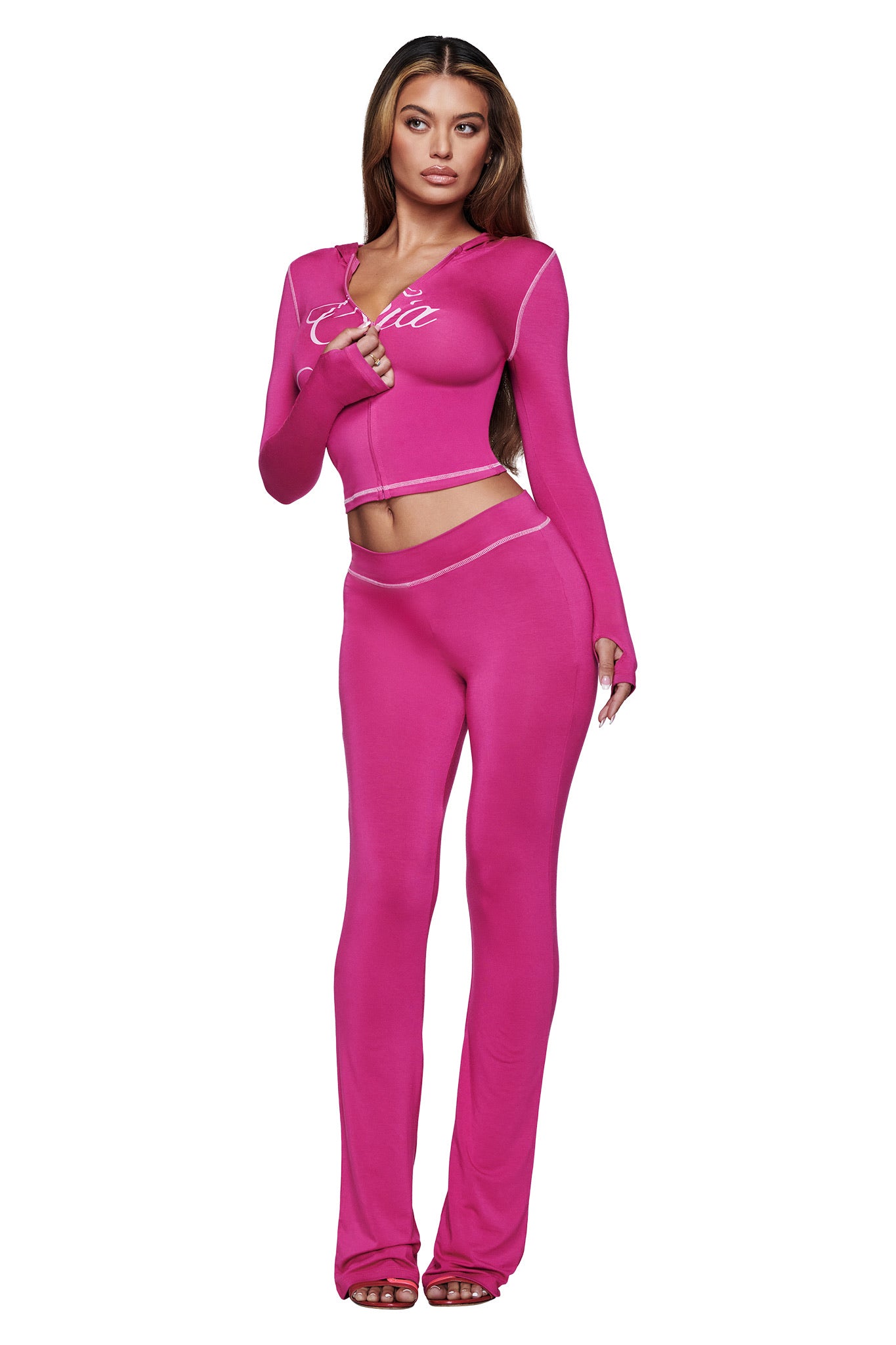 A woman with long, wavy hair faces forward against a white background, wearing I.AM.GIA’s BLARE TRACKPANT in Raspberry—a fitted two-piece set with long sleeves and flared pants—posing with one hand near her mouth.