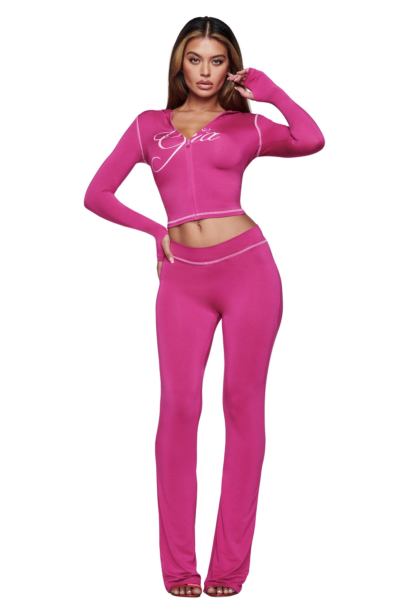 A woman faces forward in I.AM.GIA’s BLARE TRACKPANT - RASPBERRY, a vibrant pink fitted two-piece with flared pants and a long sleeve zip-up top featuring white script on the chest. She poses confidently, one hand on her hip.
