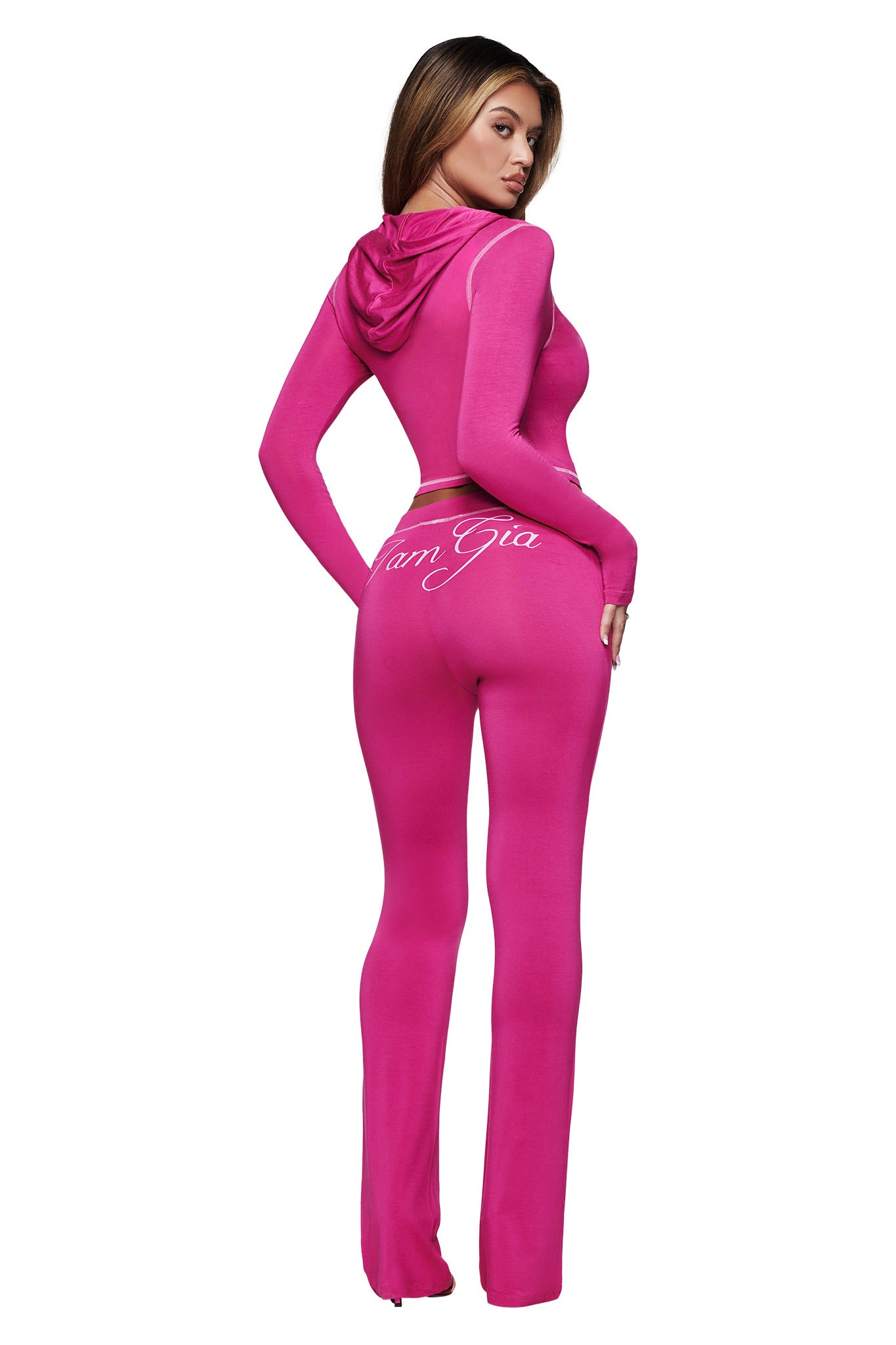 A woman poses with her back to the camera in I.AM.GIA's BLARE TRACKPANT in Raspberry, a fitted, bright pink flared pant. "I.AM.GIA" is printed in white just below the waist on the long-sleeved piece.