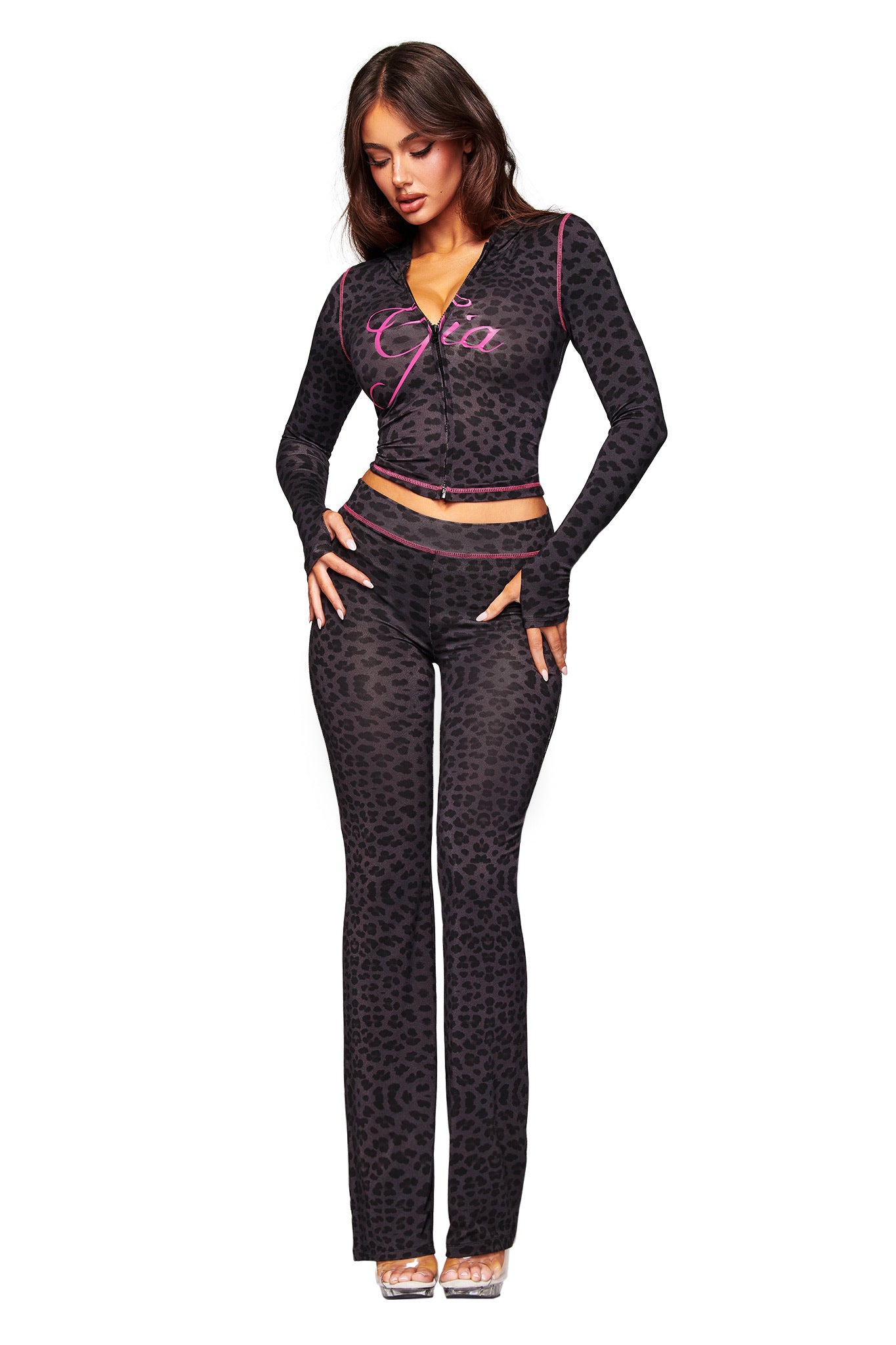 A woman models the I.AM.GIA BLARE HOODIE - PANTHER, a fitted black leopard print two-piece with long sleeves and flared pants, paired with clear heels. She poses against a white background for a chic, versatile look.