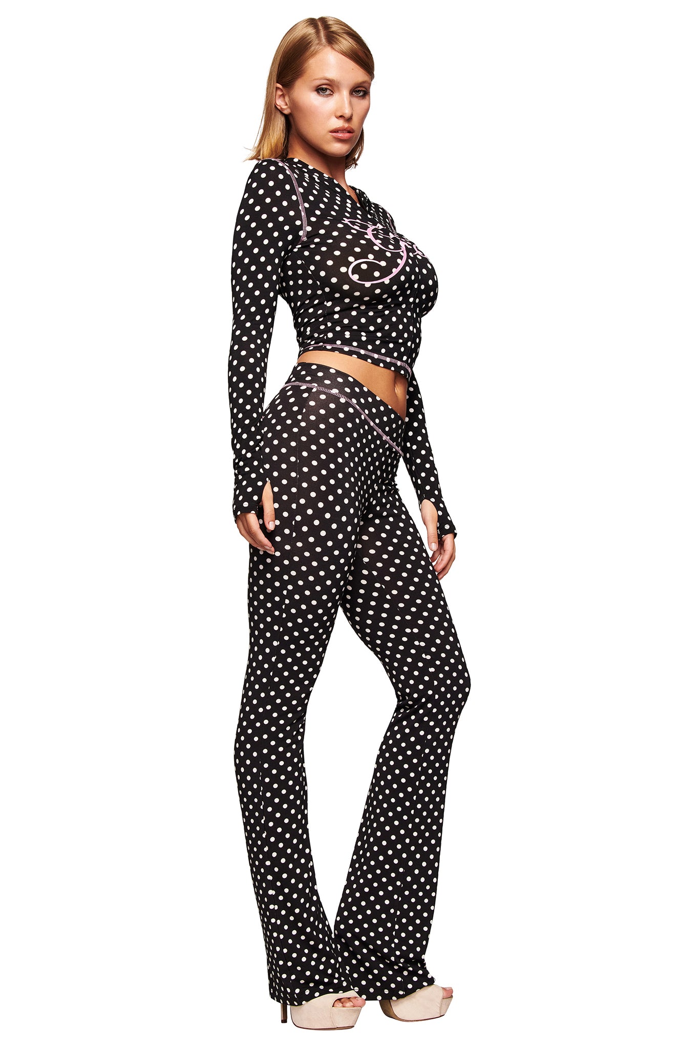 A woman poses in the I.AM.GIA BLARE HOODIE - BLACK POLKA DOT, a fitted long-sleeve top with matching flared pants in a chic polka dot print, paired with high-heeled shoes against a white background.