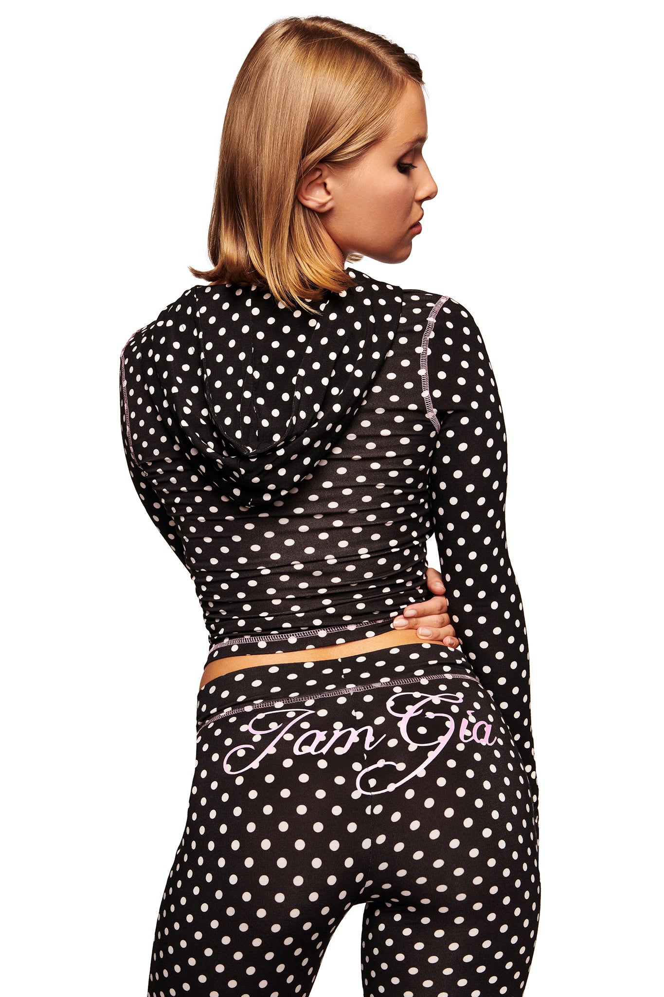 A woman with short blonde hair faces away from the camera, wearing a black hooded top and I.AM.GIA BLARE TRACKPANT - BLACK POLKA DOT, featuring a low rise waist, polka dot print, and cursive writing on the back.