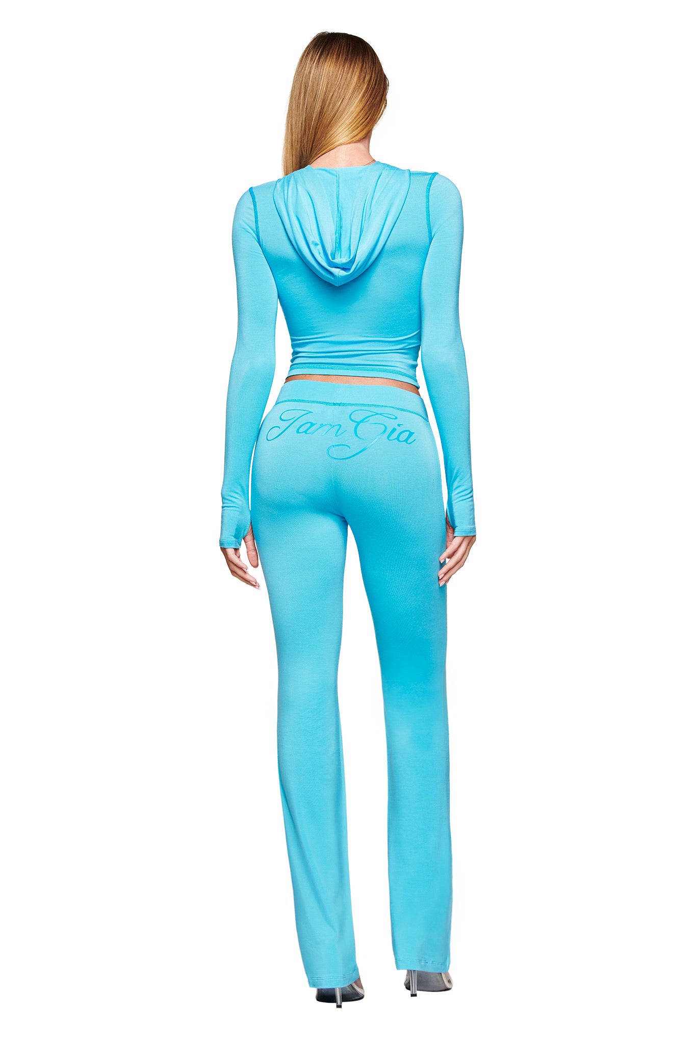 A woman with long blonde hair, seen from behind, wears the I.AM.GIA BLARE TRACKPANT in neon blue and a matching light blue hooded top, standing against a white background.