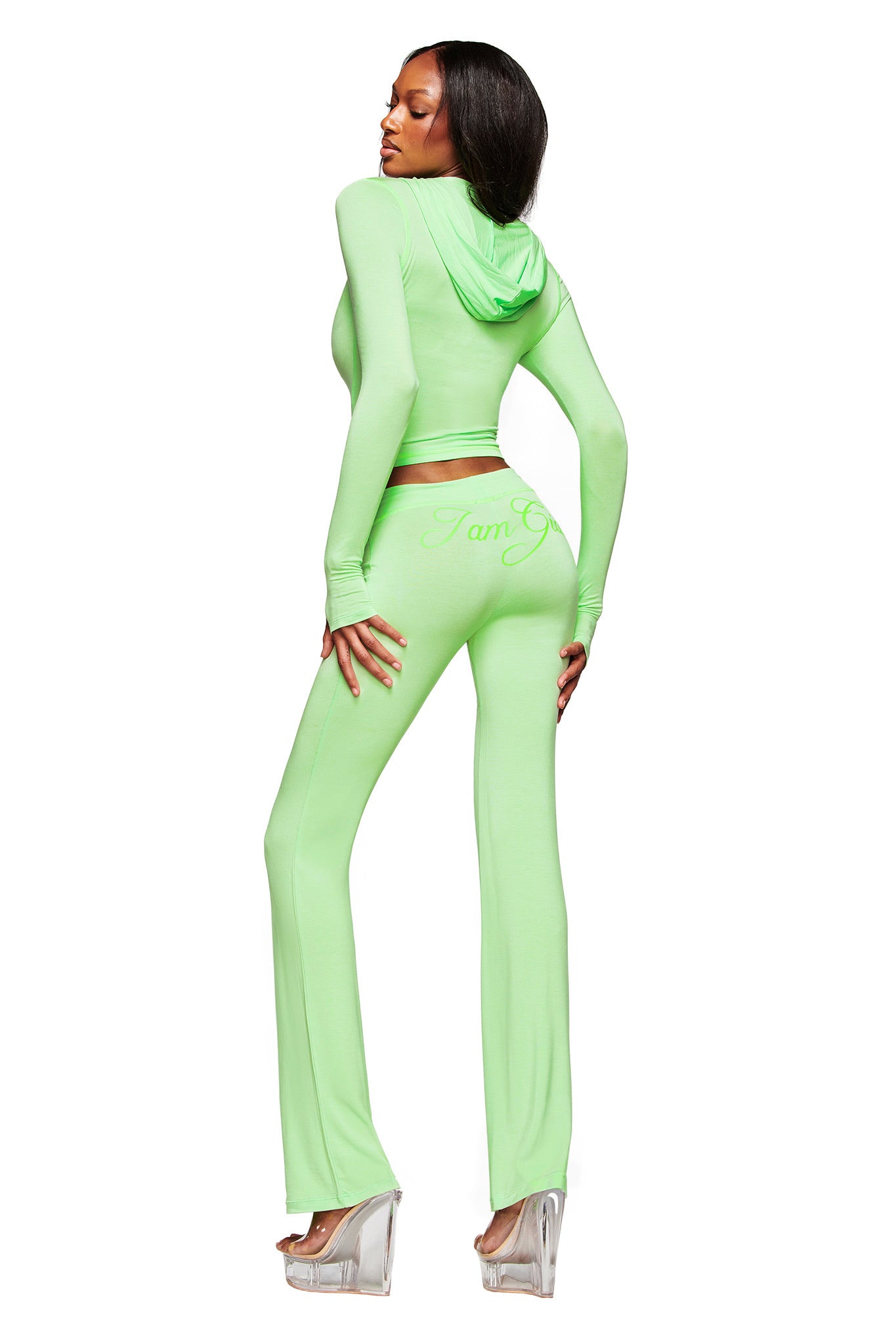 A woman models the I.AM.GIA BLARE TRACKPANT in neon green with a low-rise waist and flared legs, paired with a matching fitted hooded crop top and clear platform heels, set against a white background.