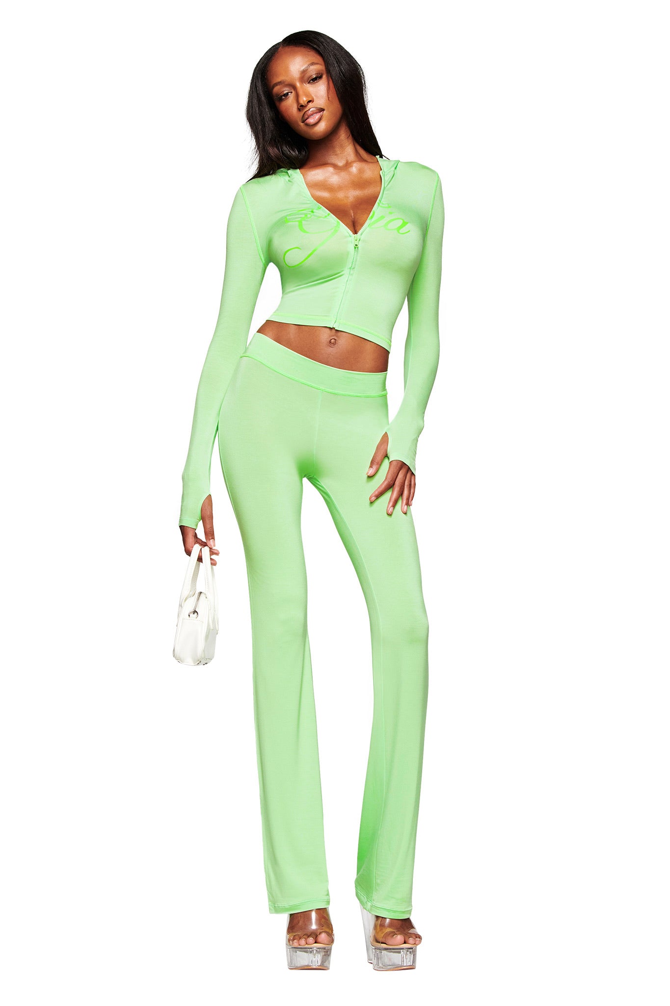 A woman models the I.AM.GIA BLARE TRACKPANT in neon green, a fitted two-piece with long sleeves and flared legs. She accessorizes with a white handbag and platform heels, showcasing the trendy low-rise waist on a plain white background.