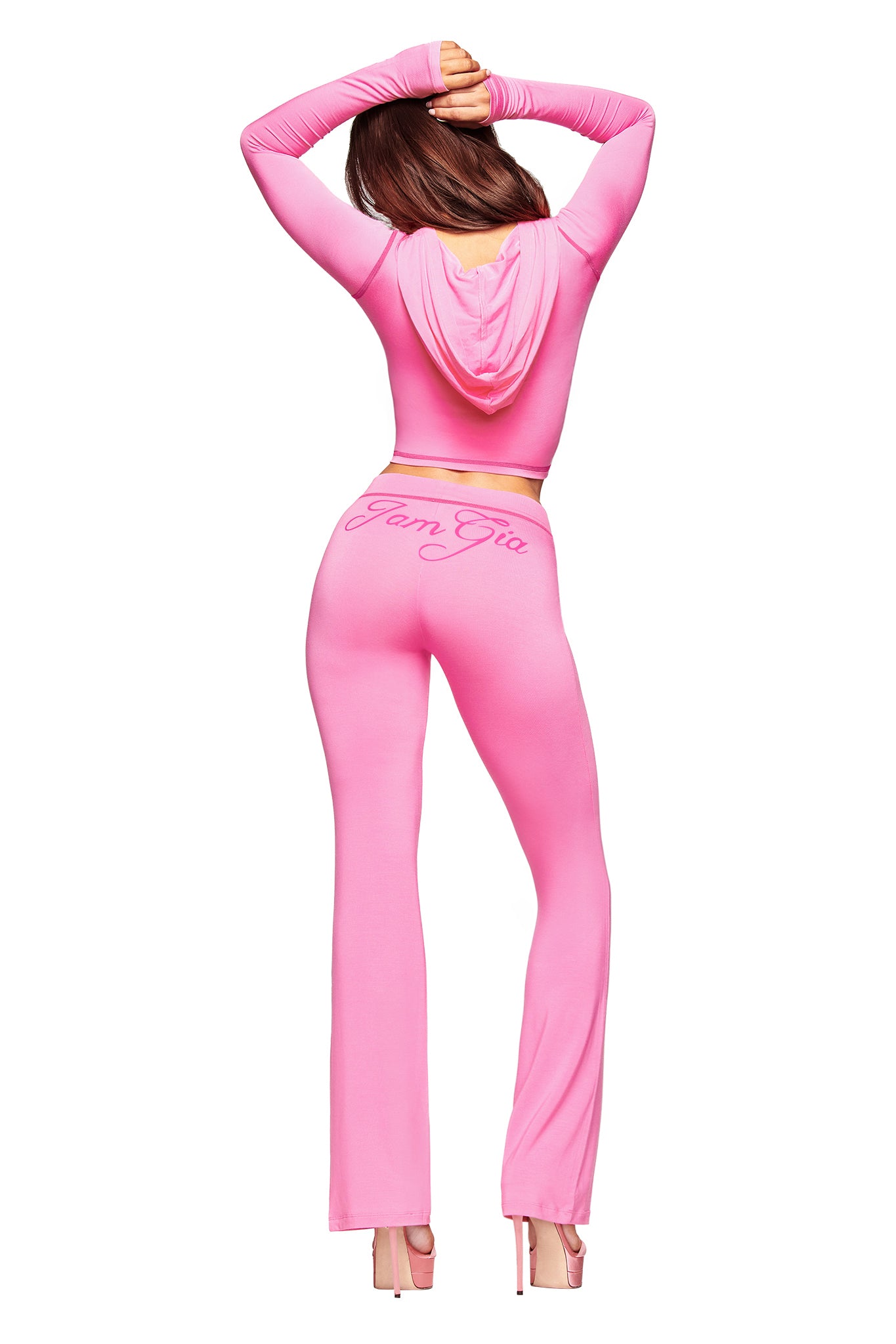 A woman poses in high heels, facing away with arms raised, wearing a fitted pink hooded crop top and the I.AM.GIA BLARE TRACKPANT in neon pink, which features the brand's logo on the back.