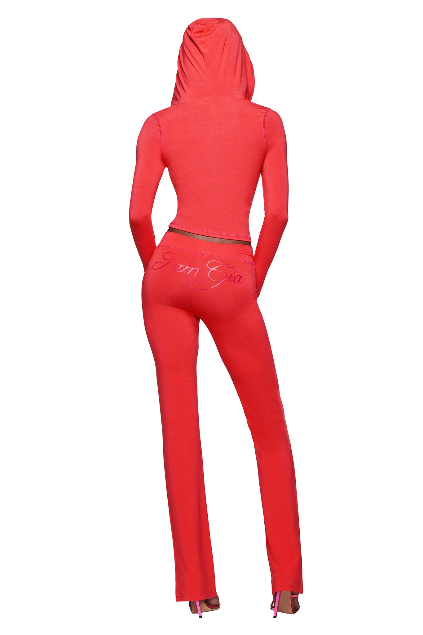 A person stands with their back to the camera, wearing a fitted, bright red hooded top, BLARE TRACKPANT - PAPAYA by I.AM.GIA with branding on the back, and pink high-heeled shoes.
