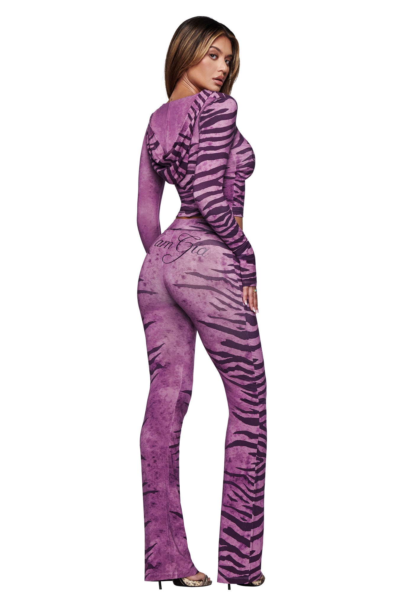 A woman stands against a white background in high heels, wearing the I.AM.GIA BLARE HOODIE jumpsuit in the Plum Wild Side print, featuring a fitted design, long sleeves, and hood, looking over her shoulder.
