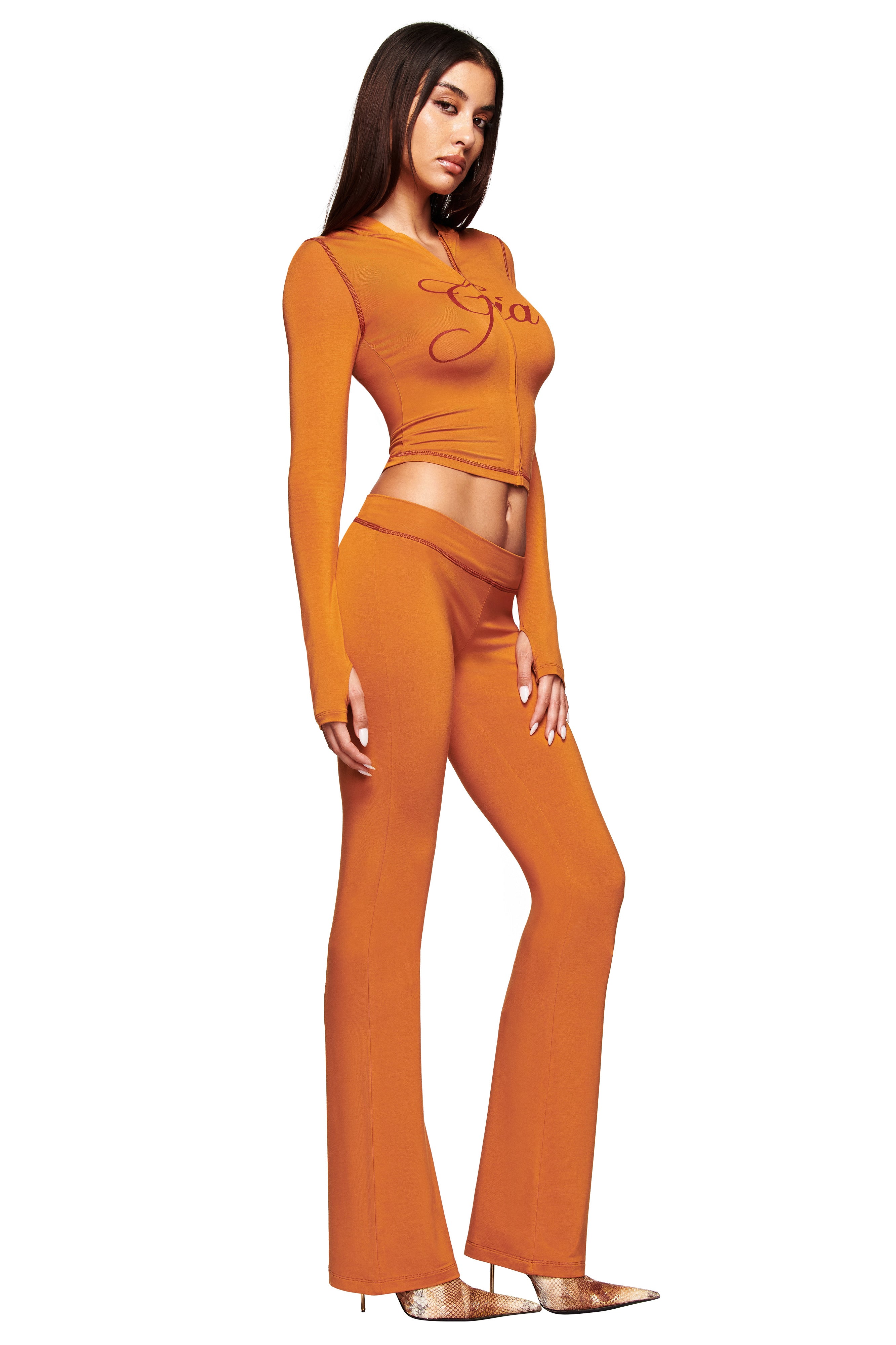 A woman stands in profile against a white background wearing the I.AM.GIA BLARE HOODIE - PUMPKIN, styled as a fitted crop top with matching high-waisted flared pants and pointed gold heels.
