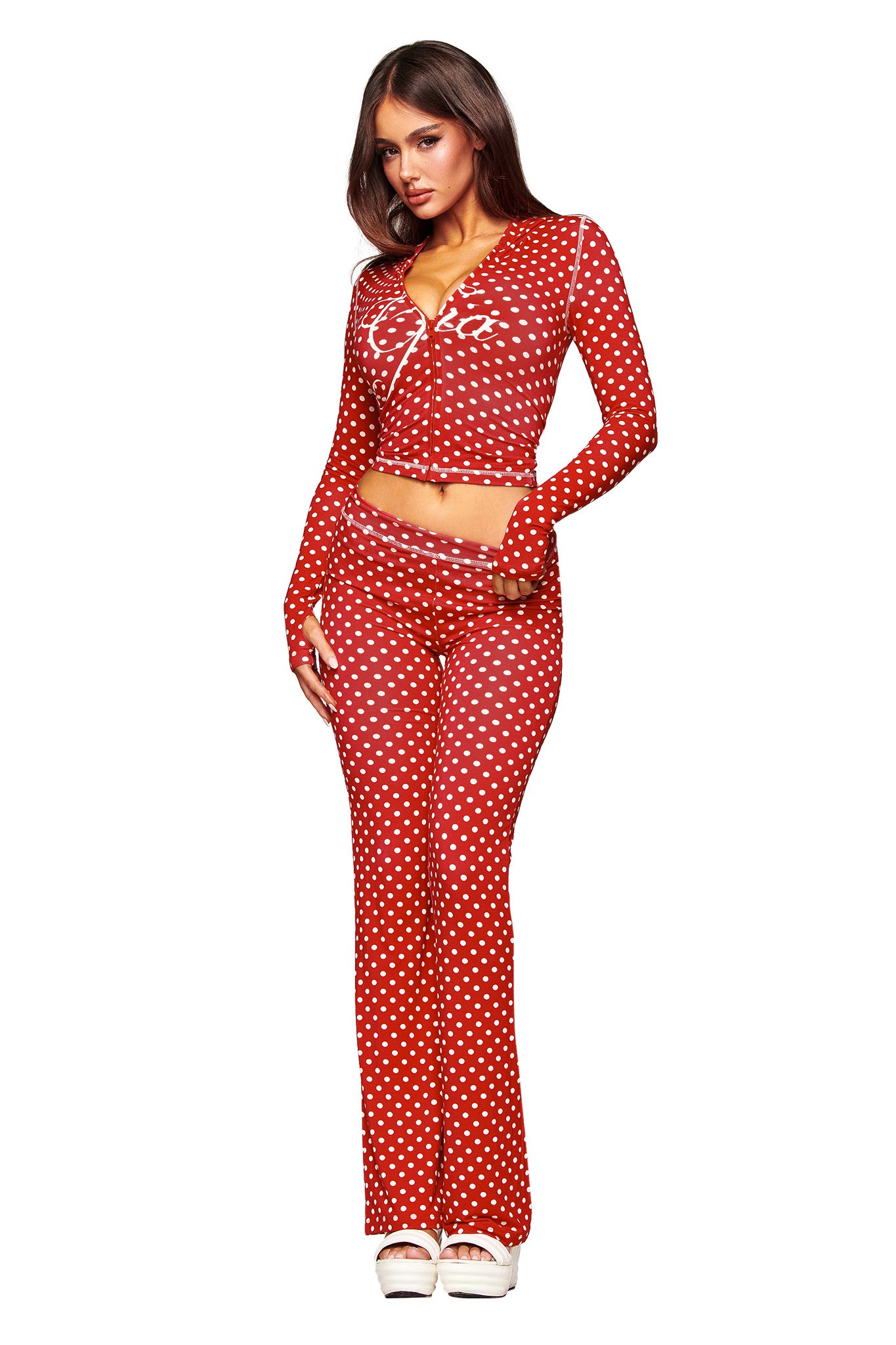 A woman with long brown hair wears I.AM.GIA’s BLARE TRACKPANT in red polka dot, styled with a matching long-sleeve V-neck crop top and white platform shoes for a chic, coordinated look.