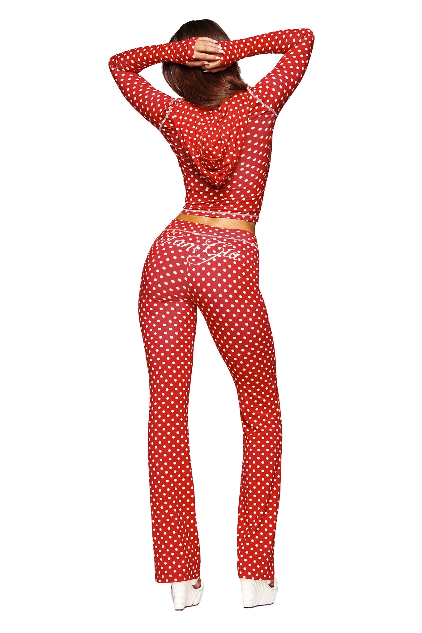 Viewed from the back, a person stands with arms raised behind their head, wearing I.AM.GIA BLARE TRACKPANT - RED POLKA DOT as part of a matching red polka dot hooded crop top set, paired with white high-heeled shoes.