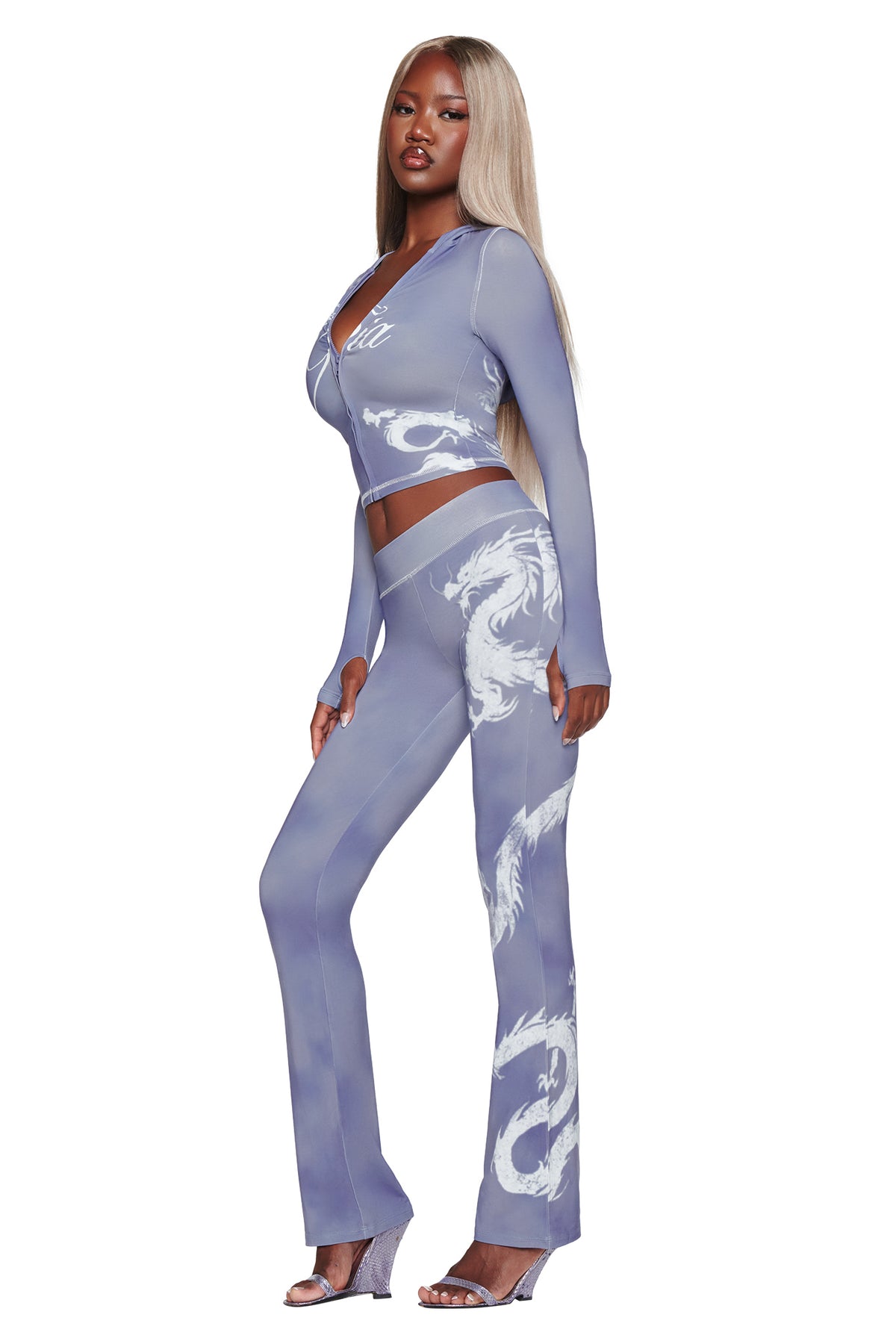 A woman stands in profile confidently wearing the I.AM.GIA BLARE TRACKPANT in a steel blue dragon print, paired with a matching fitted crop top and clear high-heeled sandals. Her long, straight blond hair completes the chic, modern look.